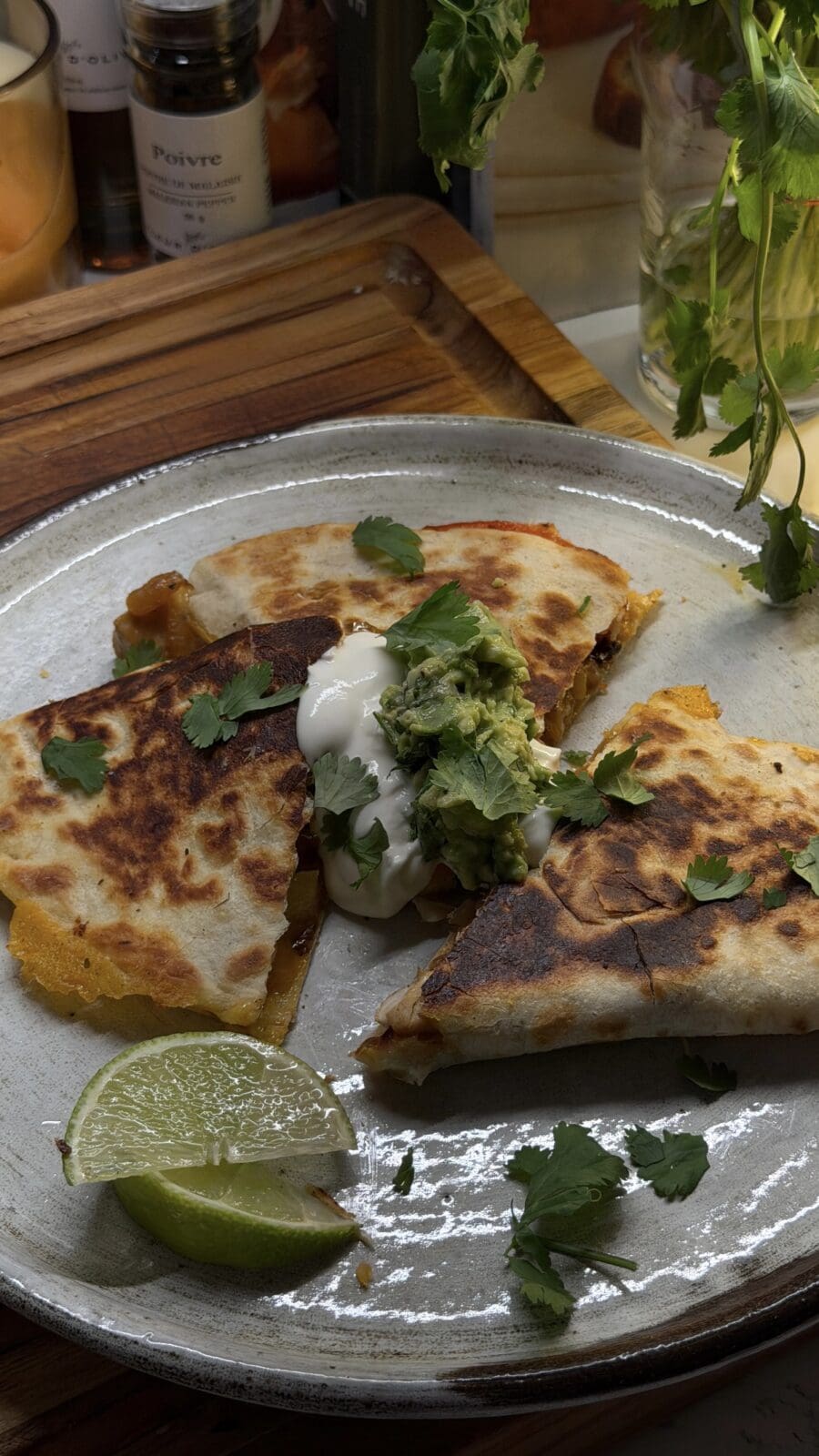 A plate with three quesadilla wedges topped with sour cream, guacamole, cilantro, and served with lime wedges.