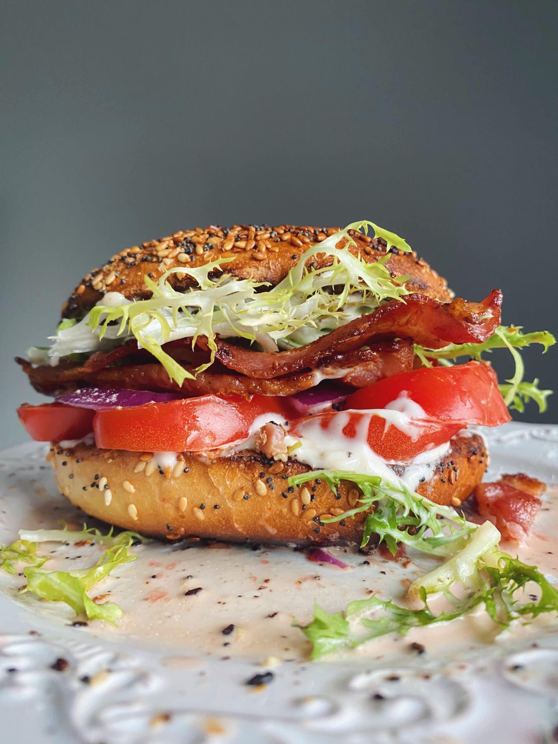 A loaded bagel sandwich with bacon, lettuce, tomato, onion, and mayonnaise sits on a white plate, with some sauce and greens spilled around it.