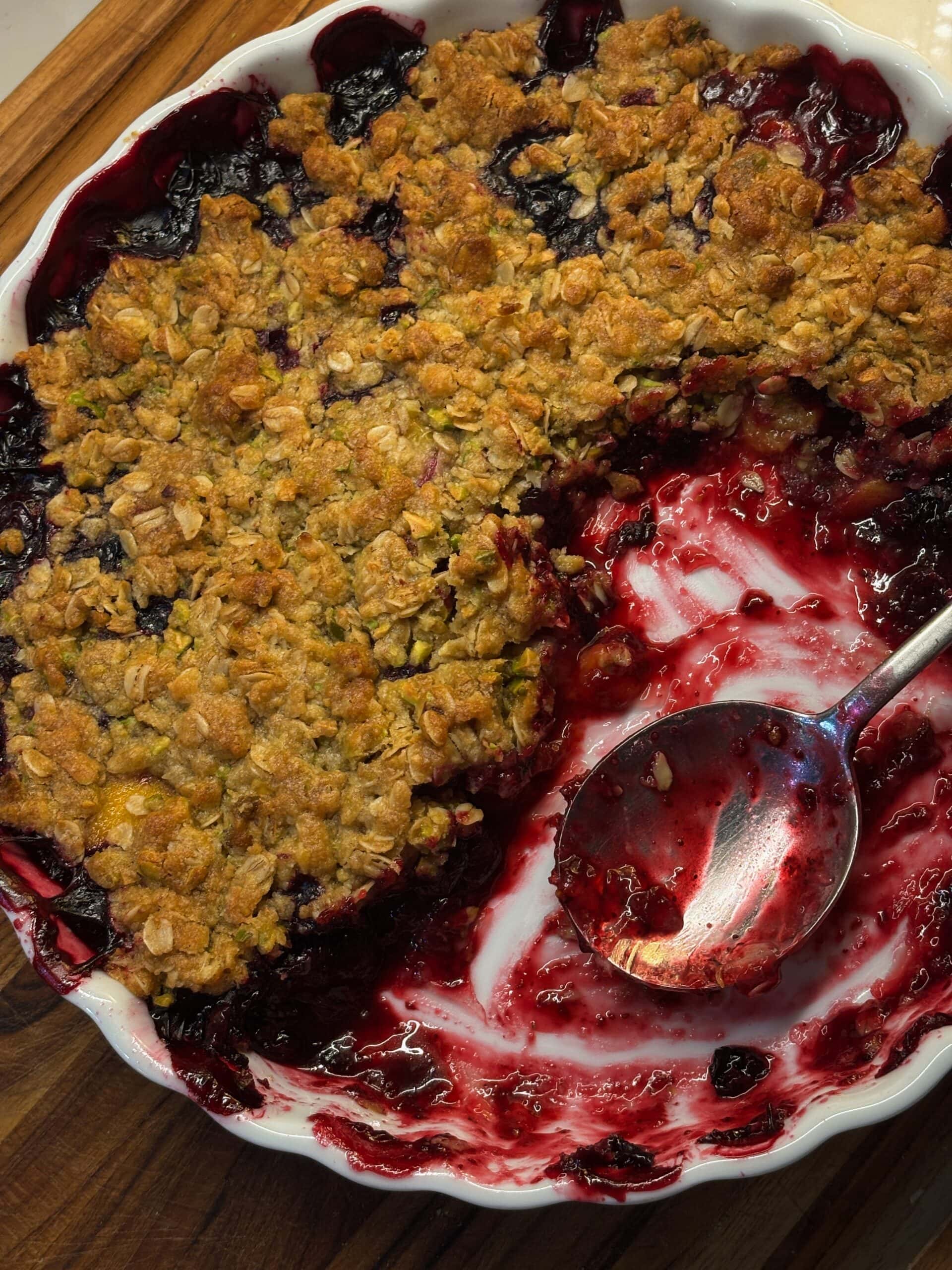 A baked berry crumble with a golden oat topping sits in a white dish; a serving has been scooped out, and a metal spoon rests in the empty section.