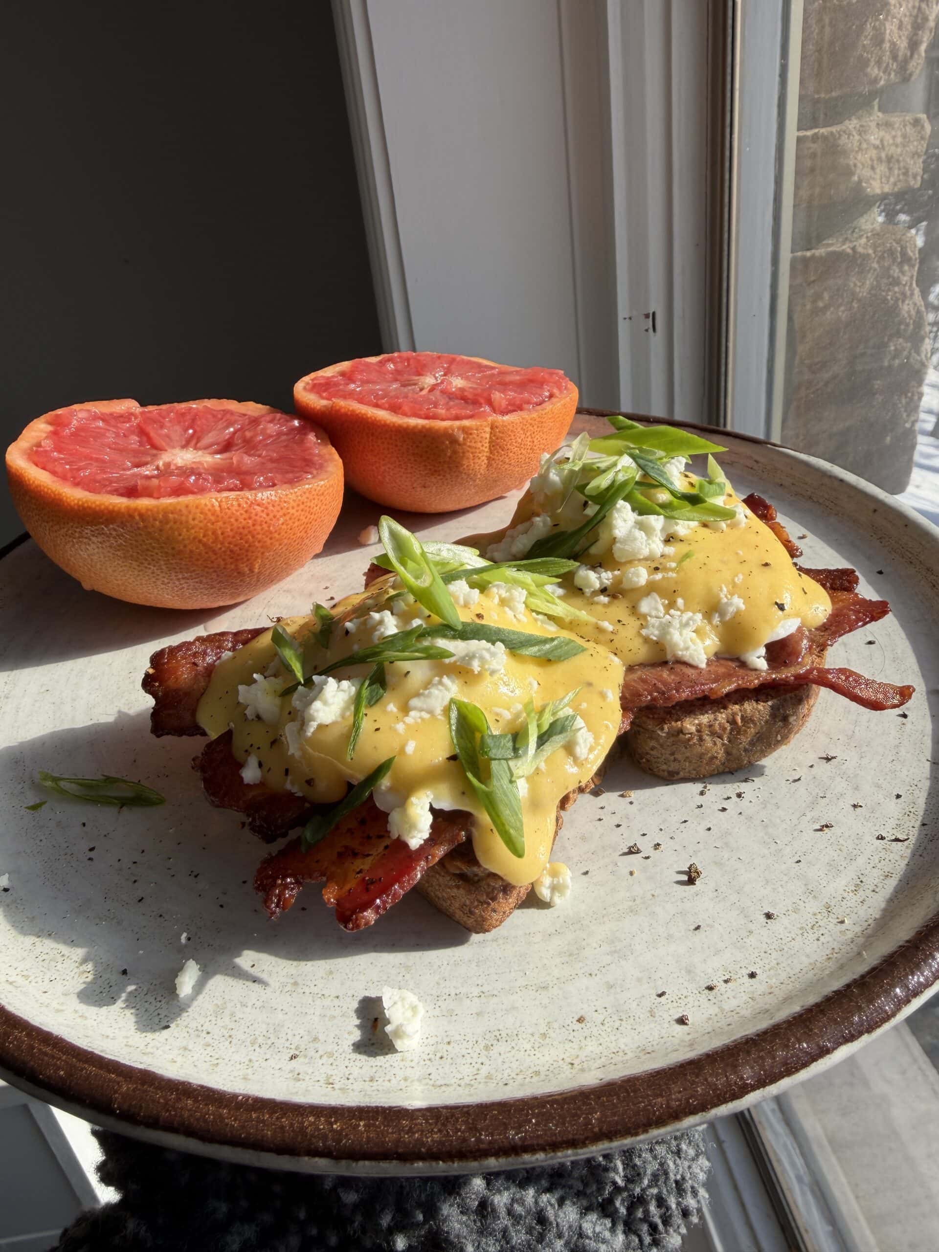 A plate with two halves of grapefruit and eggs Benedict topped with bacon, green onions, and cheese, set by a sunlit window.