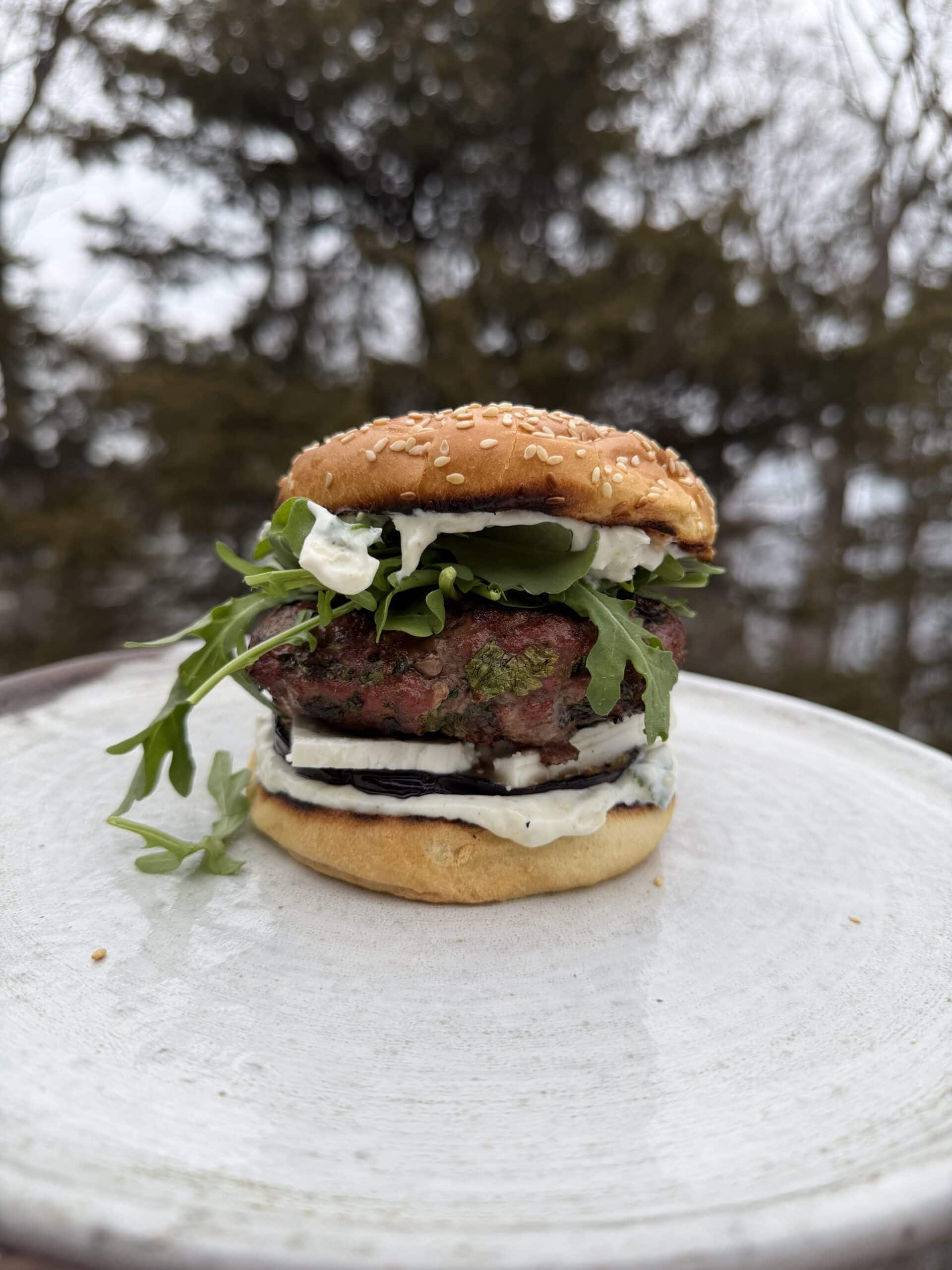 A hamburger with arugula, sliced cucumber, creamy cheese, and a sesame seed bun on a white plate with trees in the blurred background.