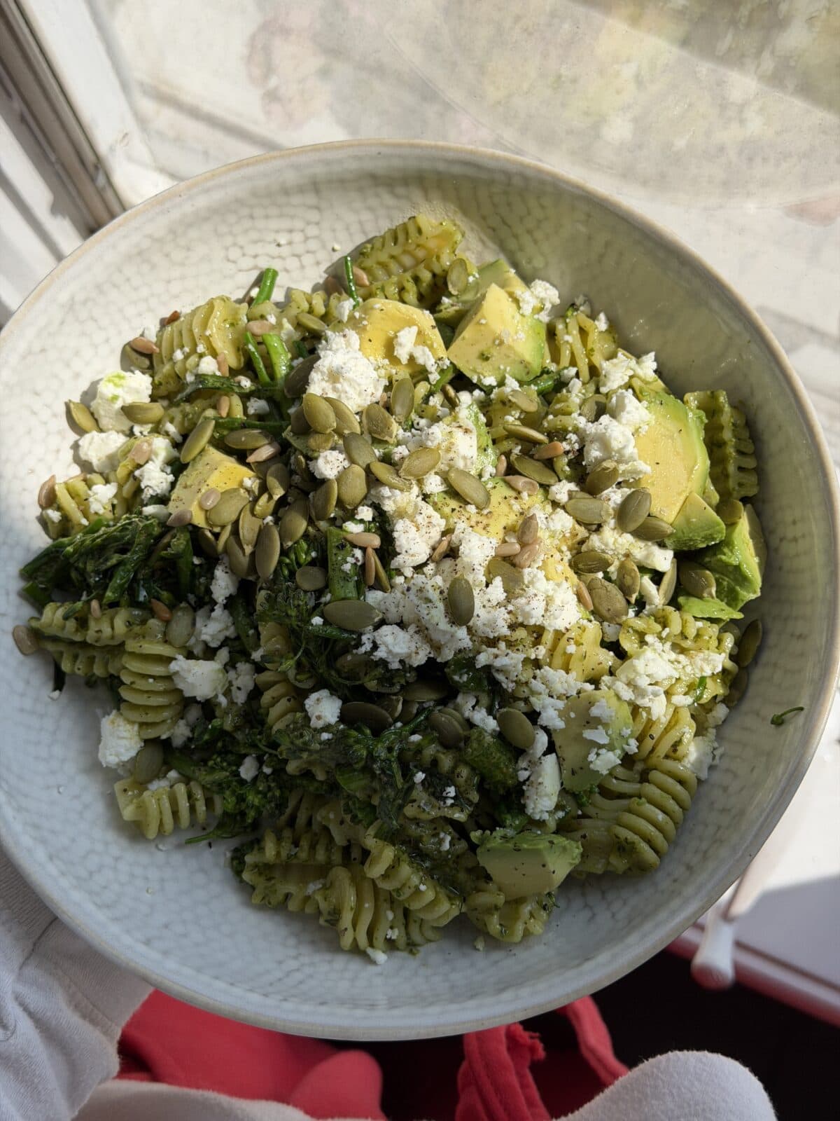 A bowl of rotini pasta topped with avocado slices, crumbled cheese, pumpkin seeds, and green vegetables, placed by a sunny window.