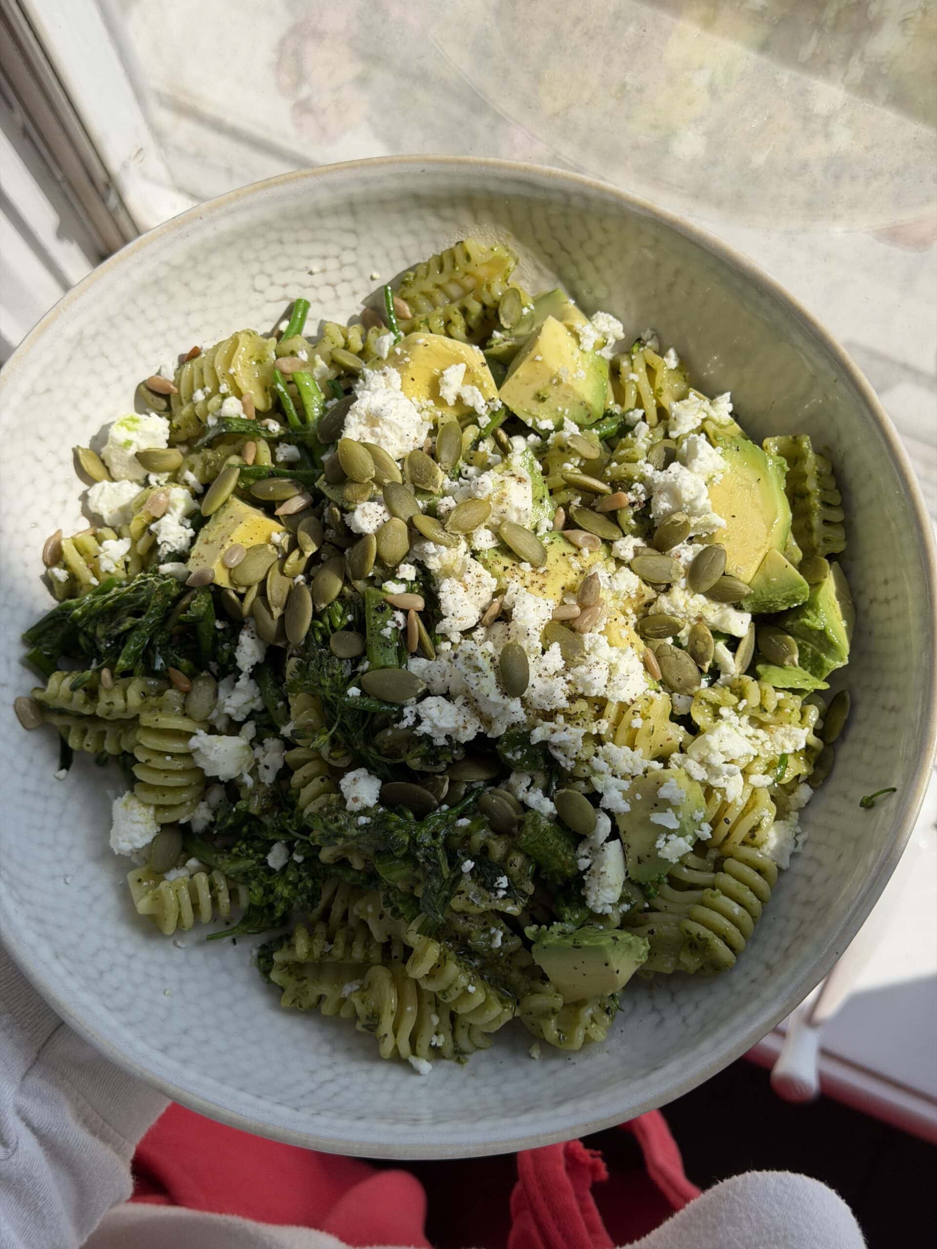 A bowl of rotini pasta topped with avocado slices, crumbled cheese, pumpkin seeds, and green vegetables, placed by a sunny window.