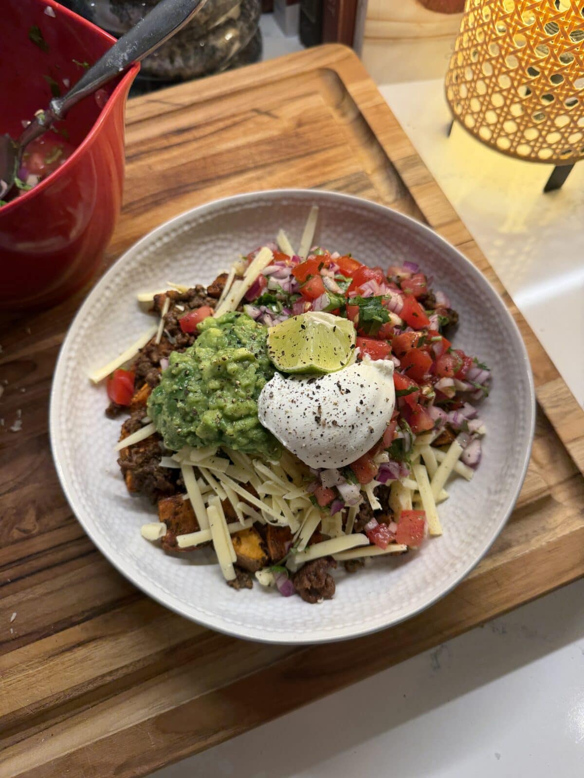 A bowl of taco salad with shredded cheese, seasoned ground beef, pico de gallo, guacamole, sour cream, and a lime wedge on top, placed on a wooden cutting board.