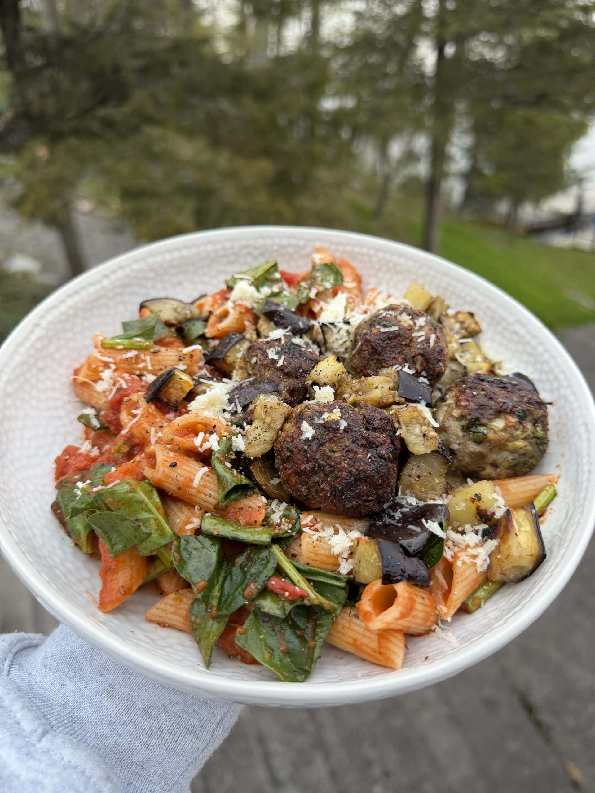 A white bowl filled with penne pasta, spinach, tomato sauce, meatballs, roasted eggplant, and grated cheese, held outdoors with trees in the background.