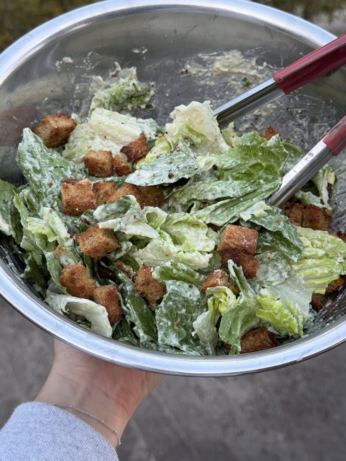 A metal bowl filled with Caesar salad, featuring romaine lettuce, croutons, and creamy dressing, being held with metal tongs.