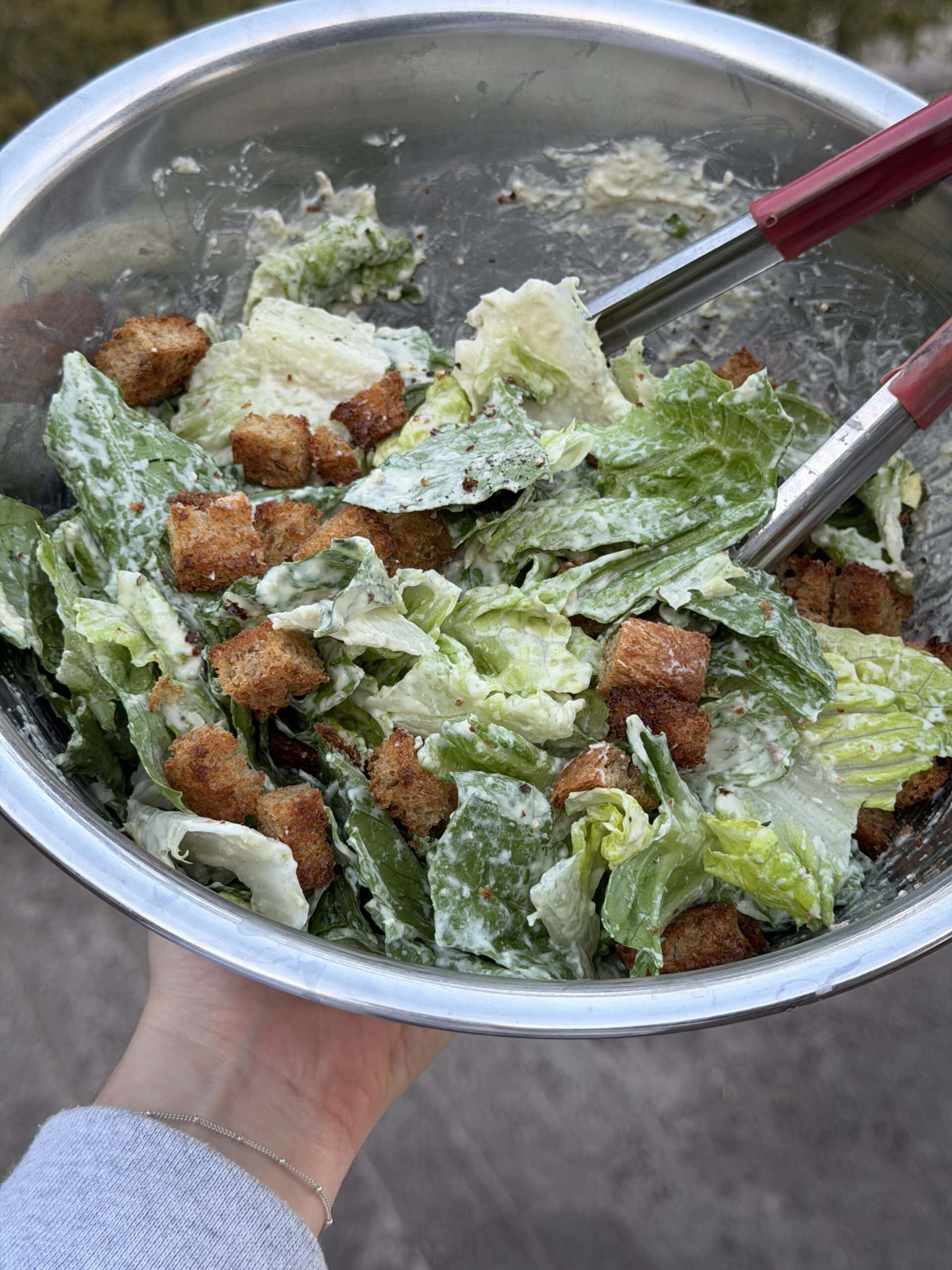 A metal bowl filled with Caesar salad, featuring romaine lettuce, croutons, and creamy dressing, being held with metal tongs.