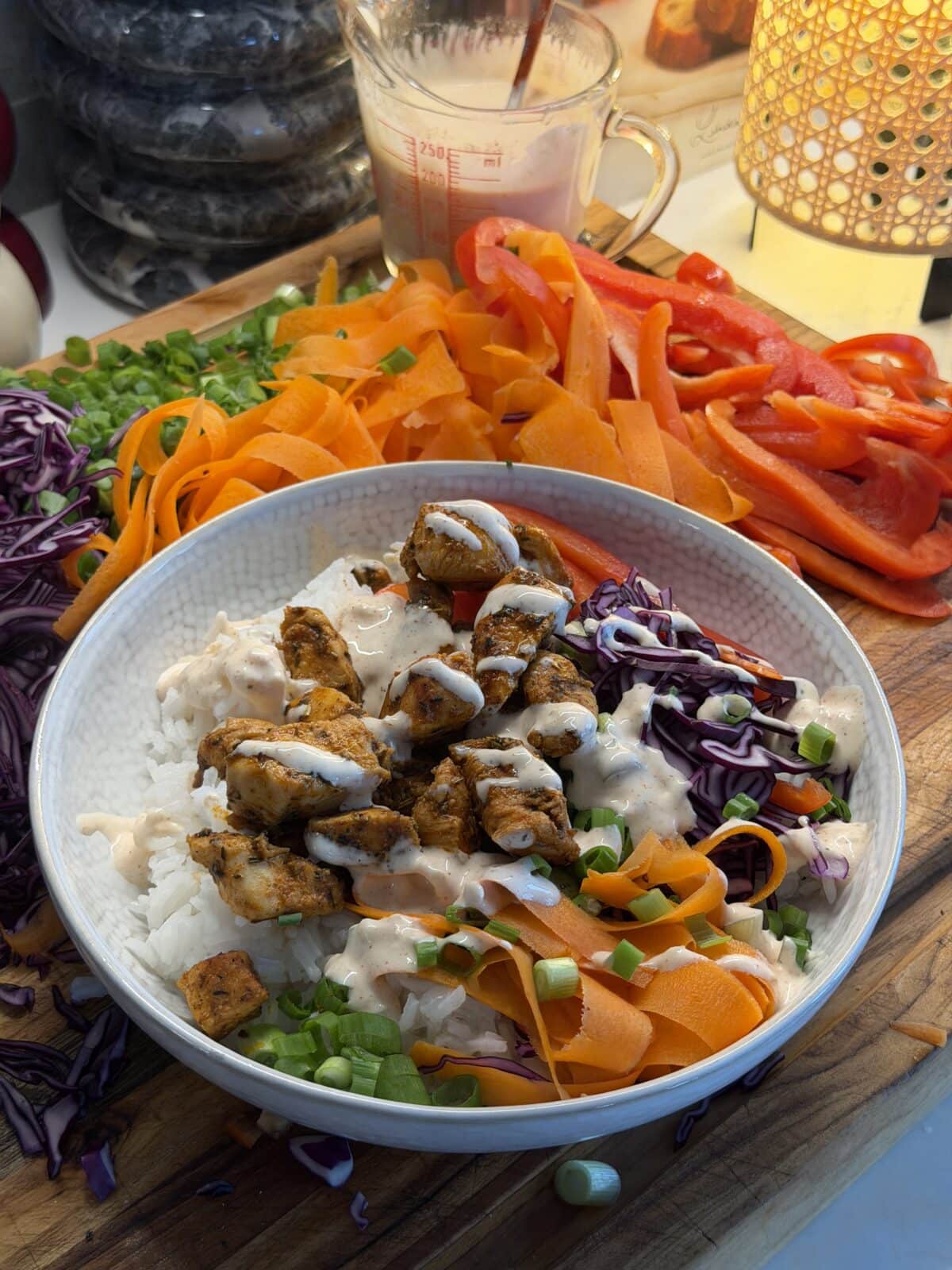 A bowl of rice topped with grilled chicken, shredded red cabbage, carrot ribbons, chopped green onions, sliced red bell pepper, and a creamy white sauce, surrounded by fresh vegetables.