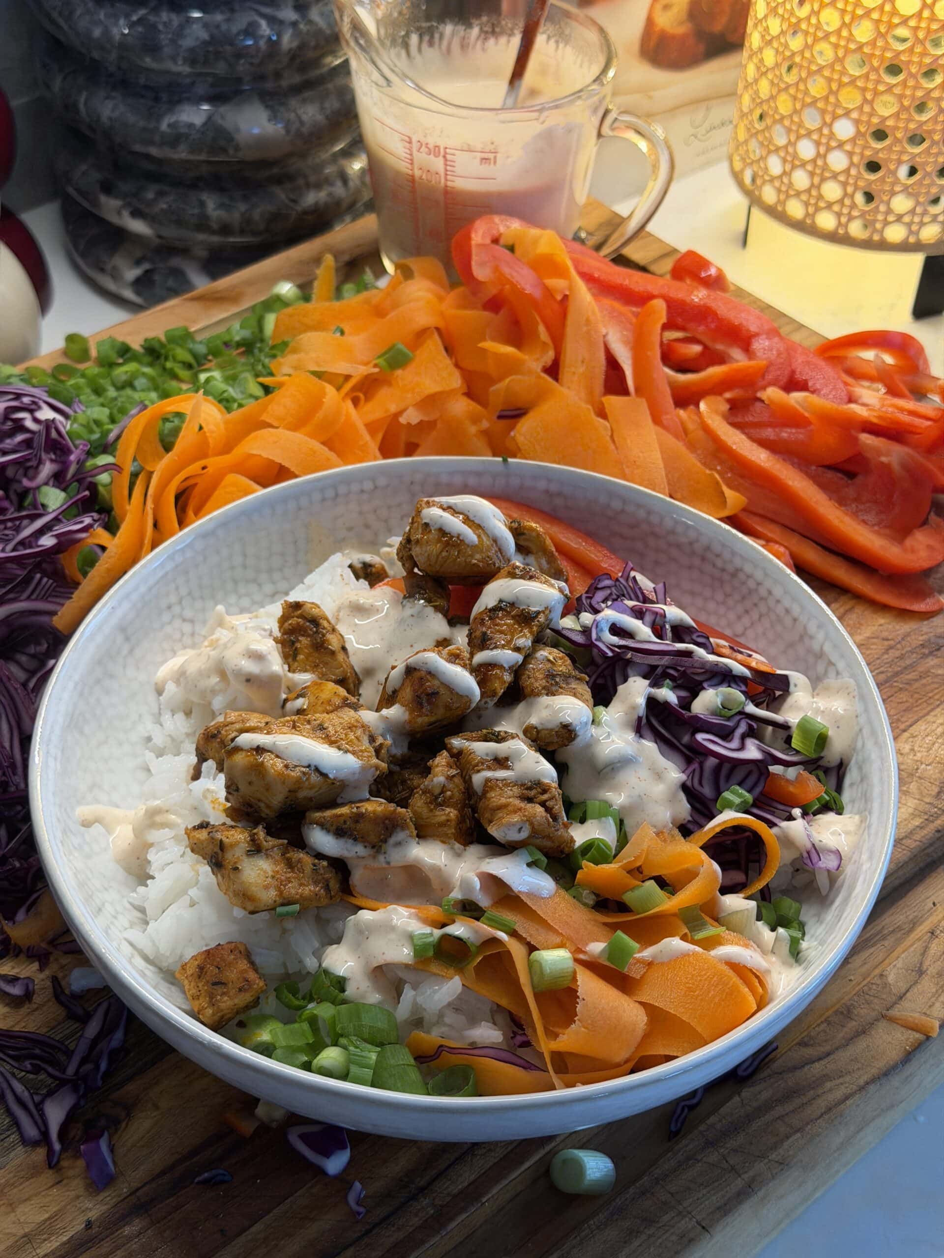A bowl of rice topped with grilled chicken, shredded red cabbage, carrot ribbons, chopped green onions, sliced red bell pepper, and a creamy white sauce, surrounded by fresh vegetables.