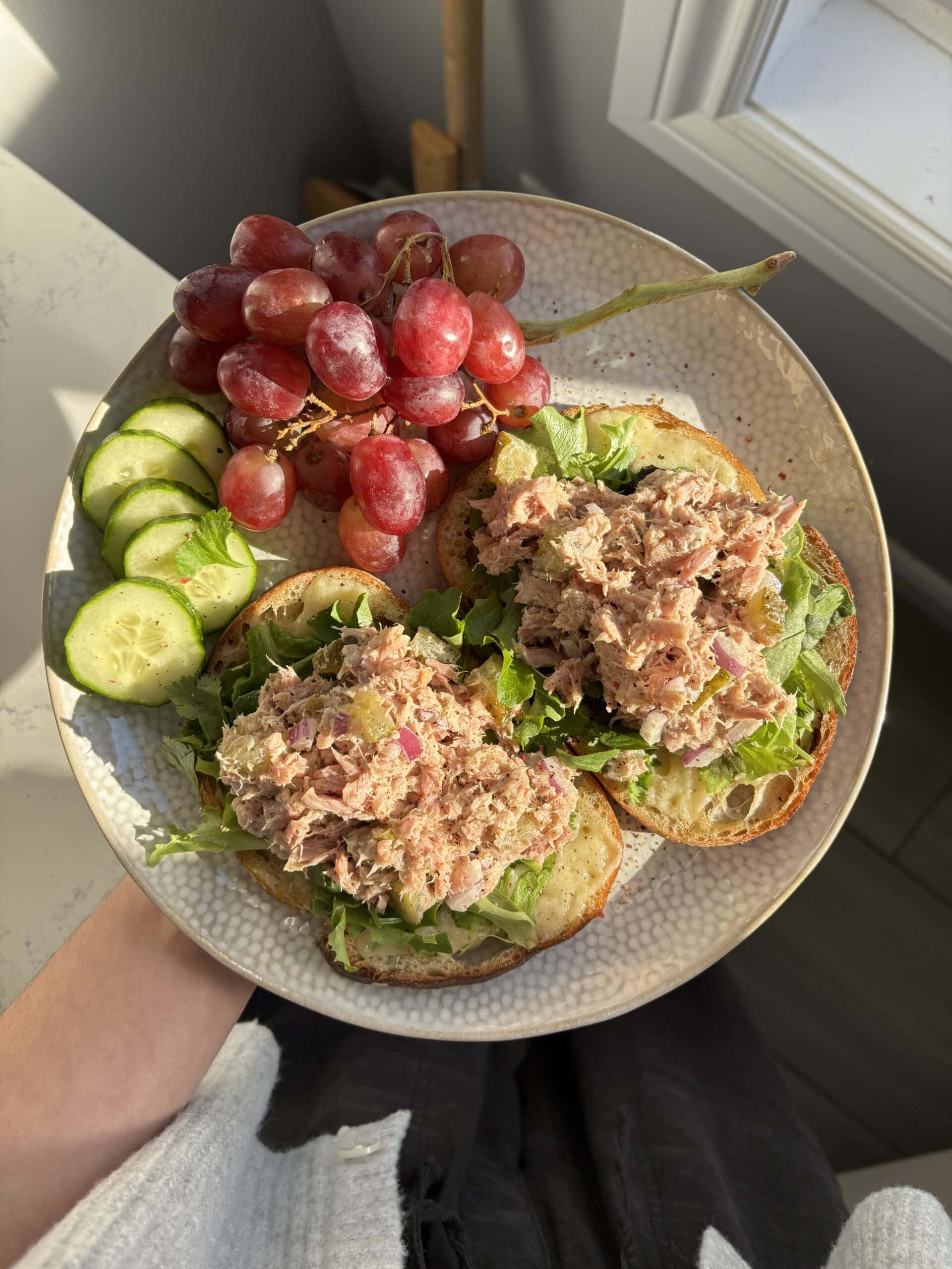 A plate with two open-faced tuna salad sandwiches on lettuce, sliced cucumbers, and a bunch of red grapes, held near a sunlit window.