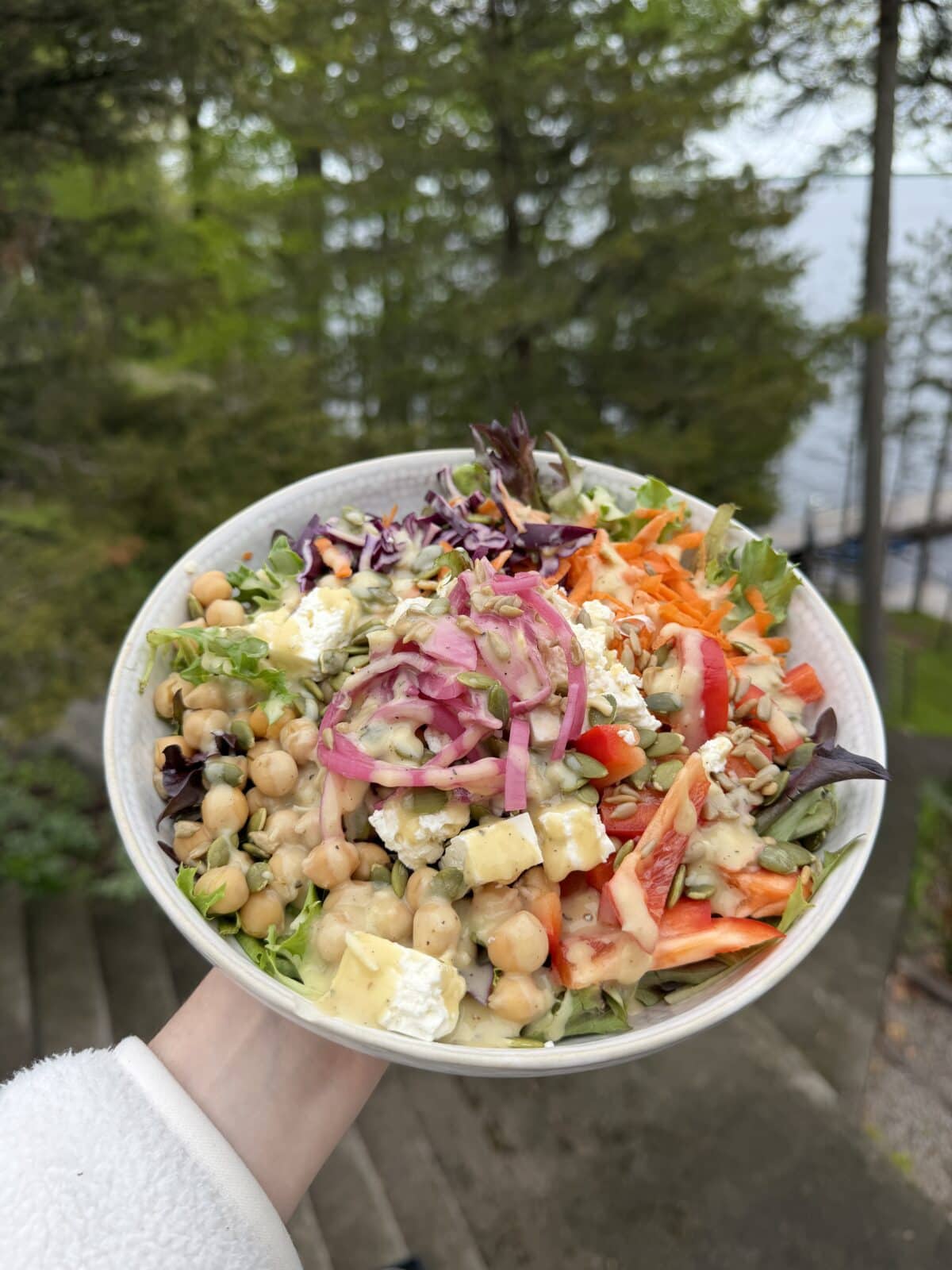 A white bowl filled with a salad containing chickpeas, feta cheese, shredded carrots, red onion, bell peppers, and seeds, drizzled with dressing.