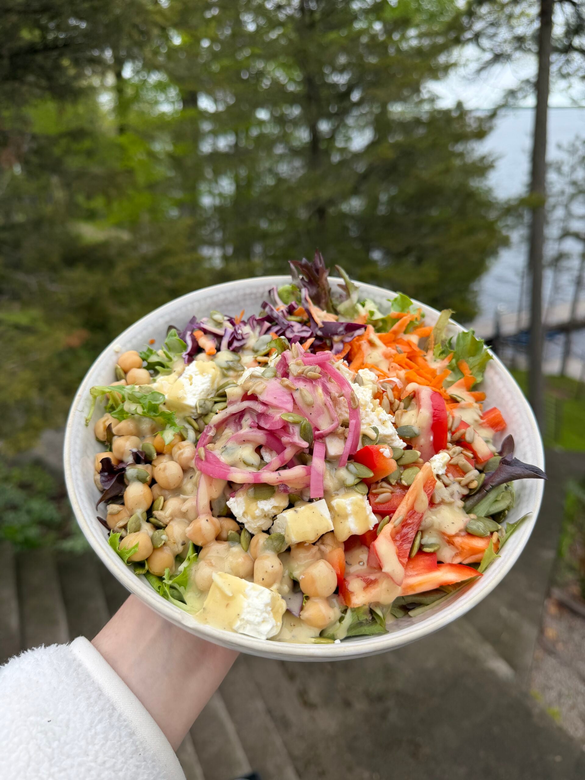 A white bowl filled with a salad containing chickpeas, feta cheese, shredded carrots, red onion, bell peppers, and seeds, drizzled with dressing.