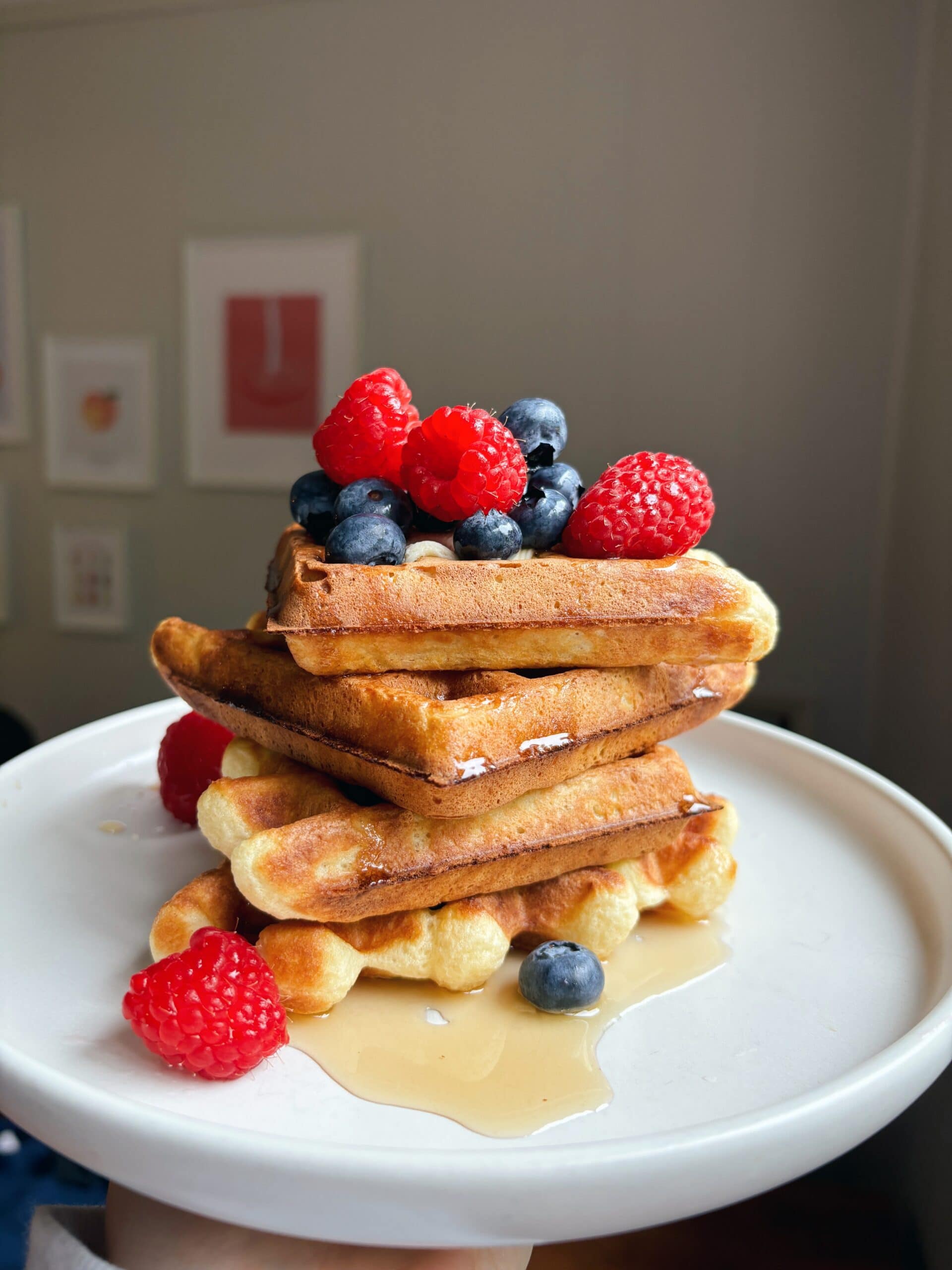 A stack of waffles topped with raspberries, blueberries, and syrup on a white plate, with framed art visible in the background.