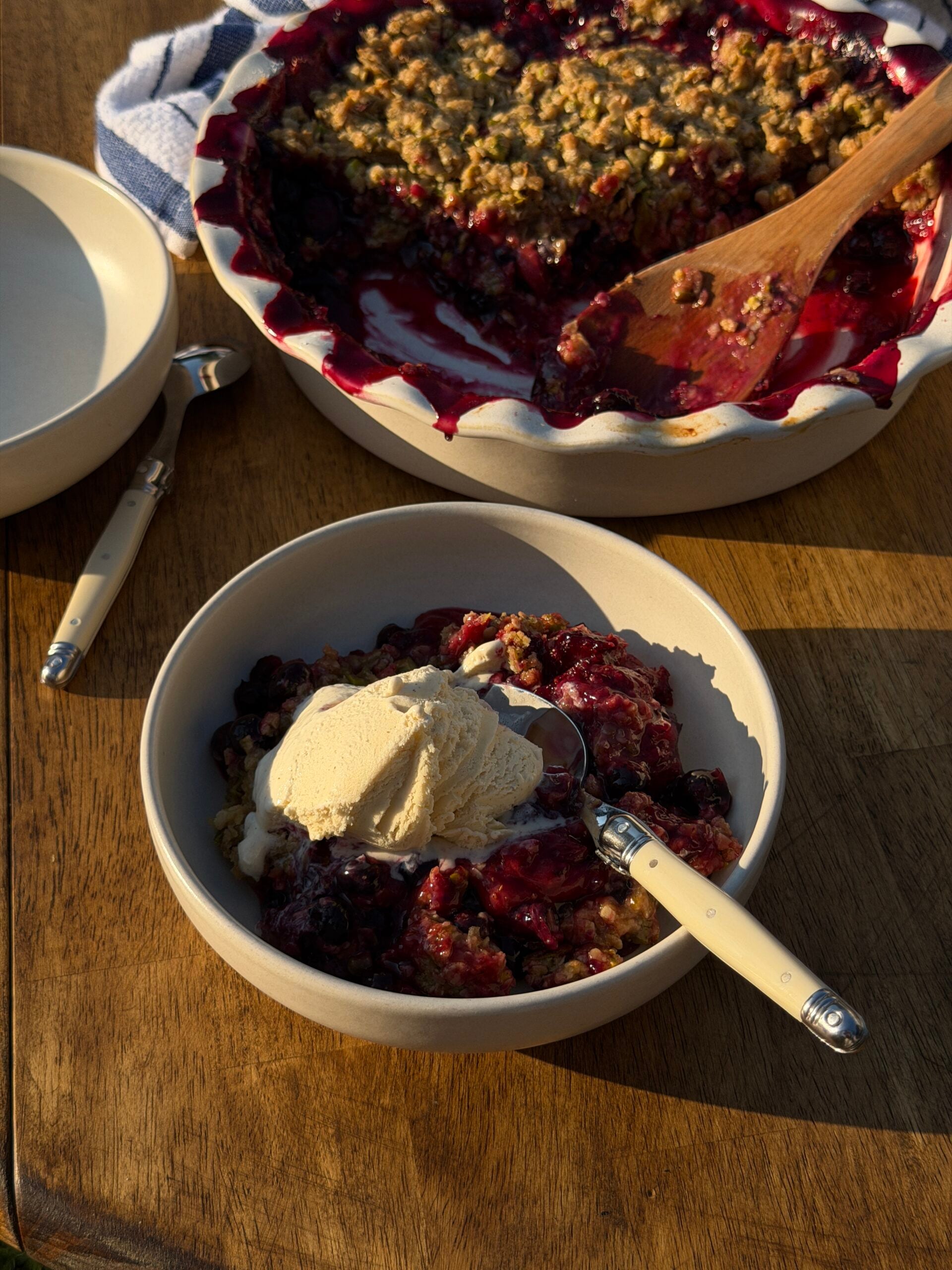 A bowl of berry crumble topped with vanilla ice cream sits on a wooden table next to a serving dish of crumble and empty plates.