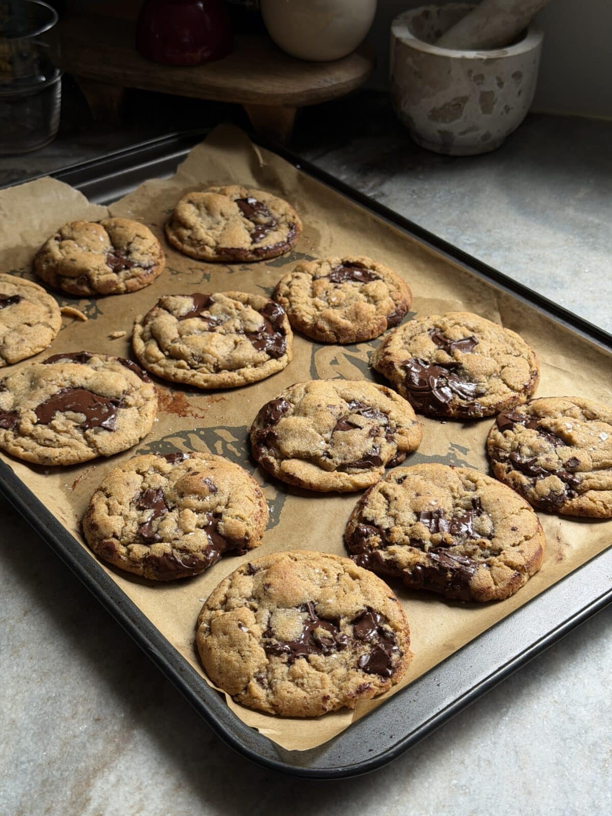 A baking tray lined with parchment paper holds twelve freshly baked chocolate chip cookies cooling on a countertop.