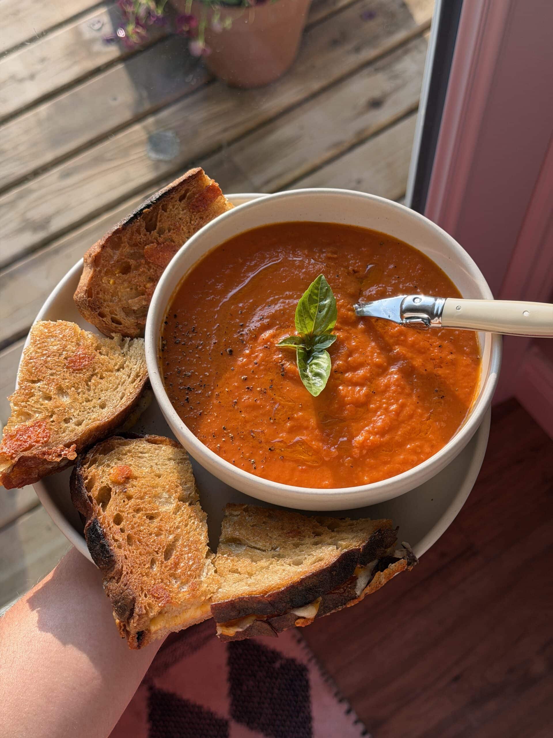A bowl of tomato soup garnished with basil, served with toasted bread slices on a plate, and a spoon resting in the soup.