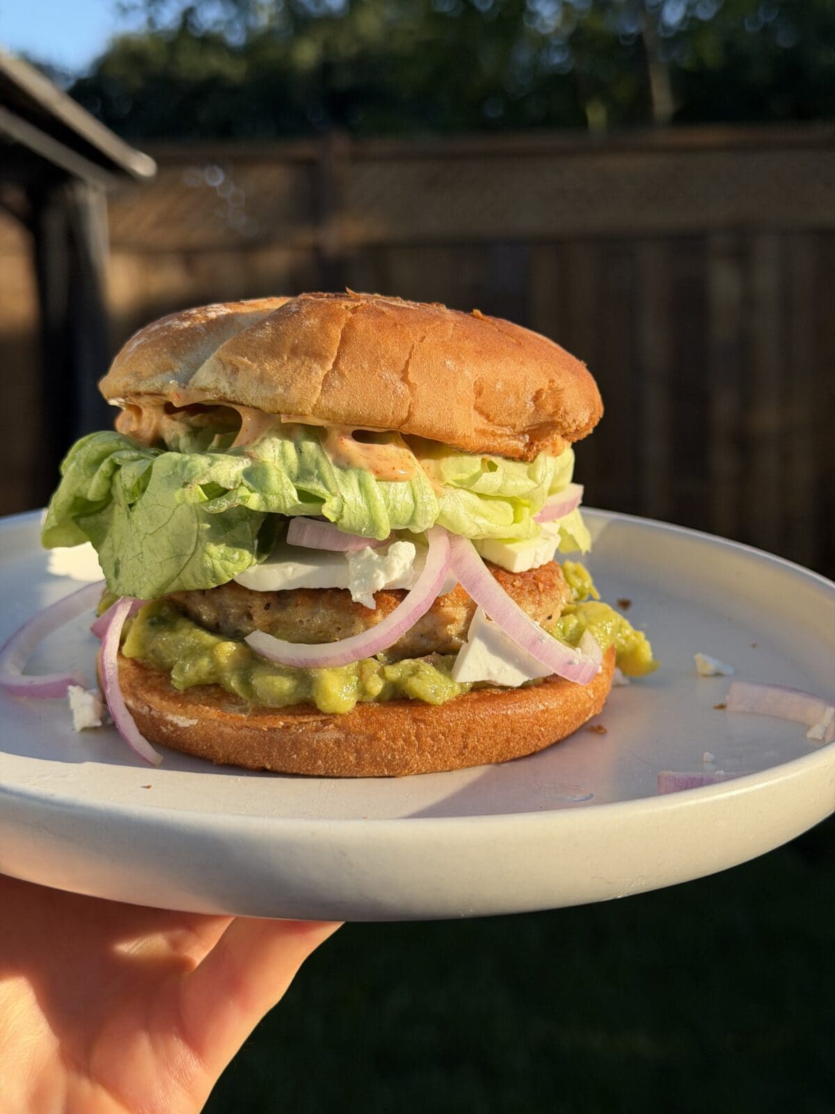 A hand holds a plate with a sandwich featuring lettuce, guacamole, sliced onions, white cheese, and sauce in a toasted bun. A wooden fence and greenery are in the background.