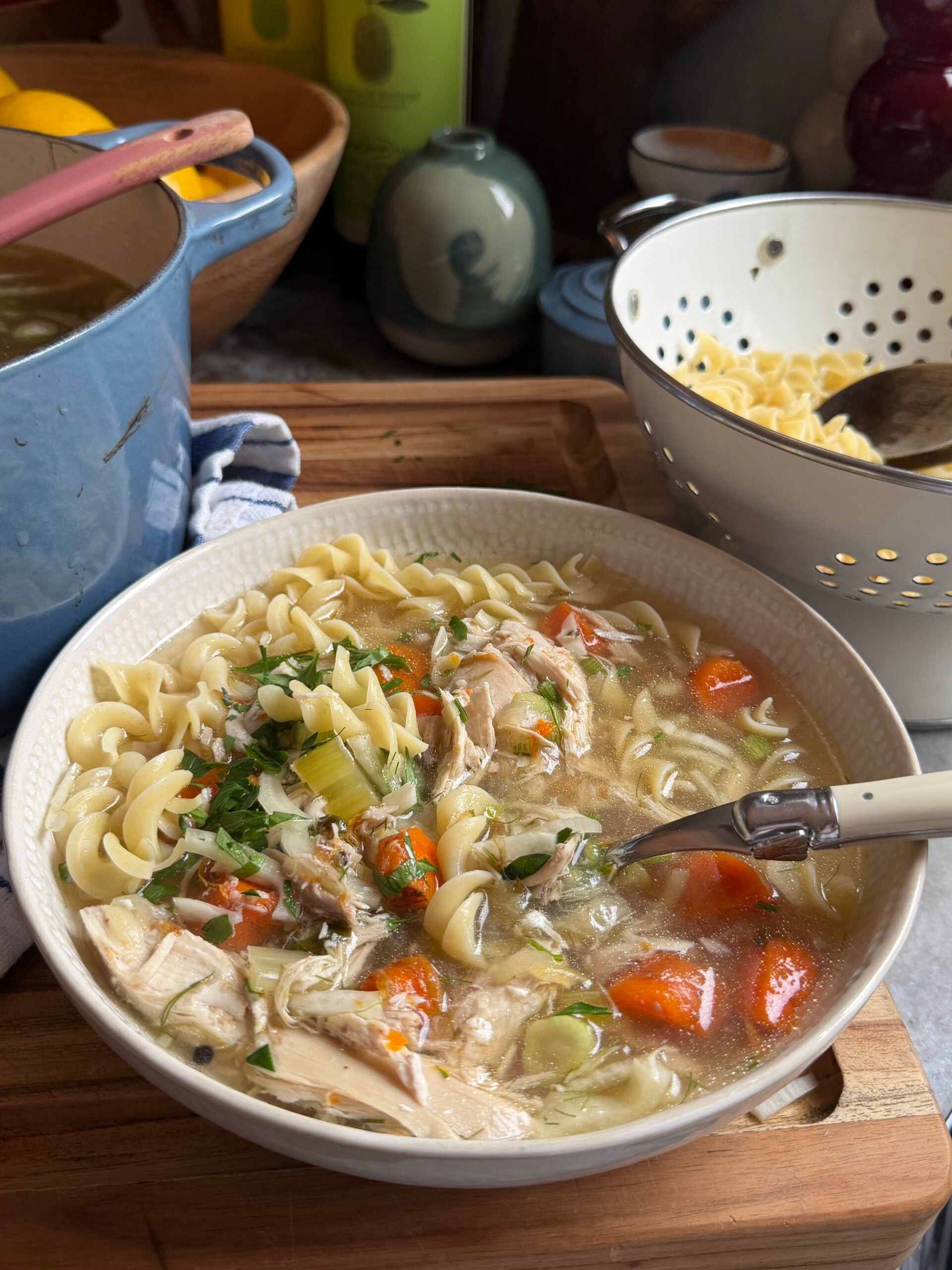 A bowl of chicken noodle soup with rotini pasta, carrots, and herbs next to a pot and a colander of cooked noodles on a wooden surface.