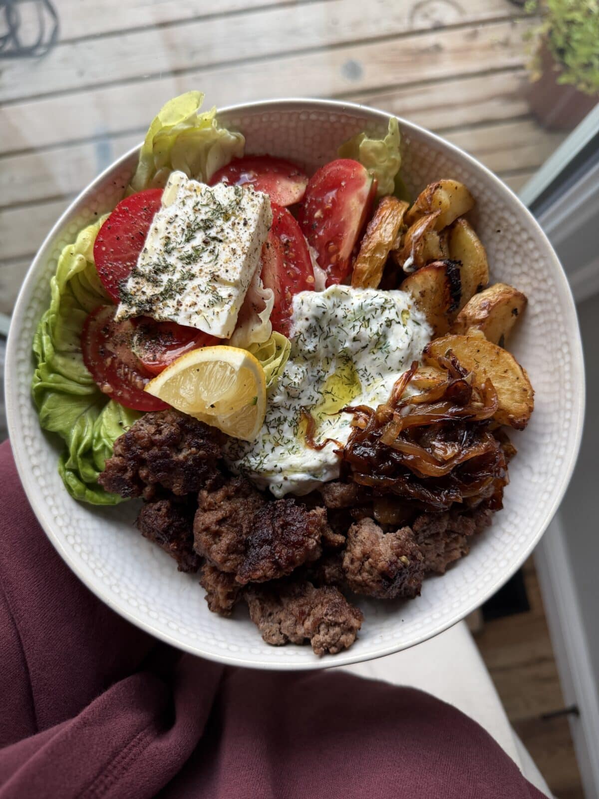 A Greek bowl featuring lettuce, tomato, feta, roasted potatoes, grilled beef, caramelized onions, tzatziki, olive oil, herbs, and a lemon wedge is beautifully set near a window.