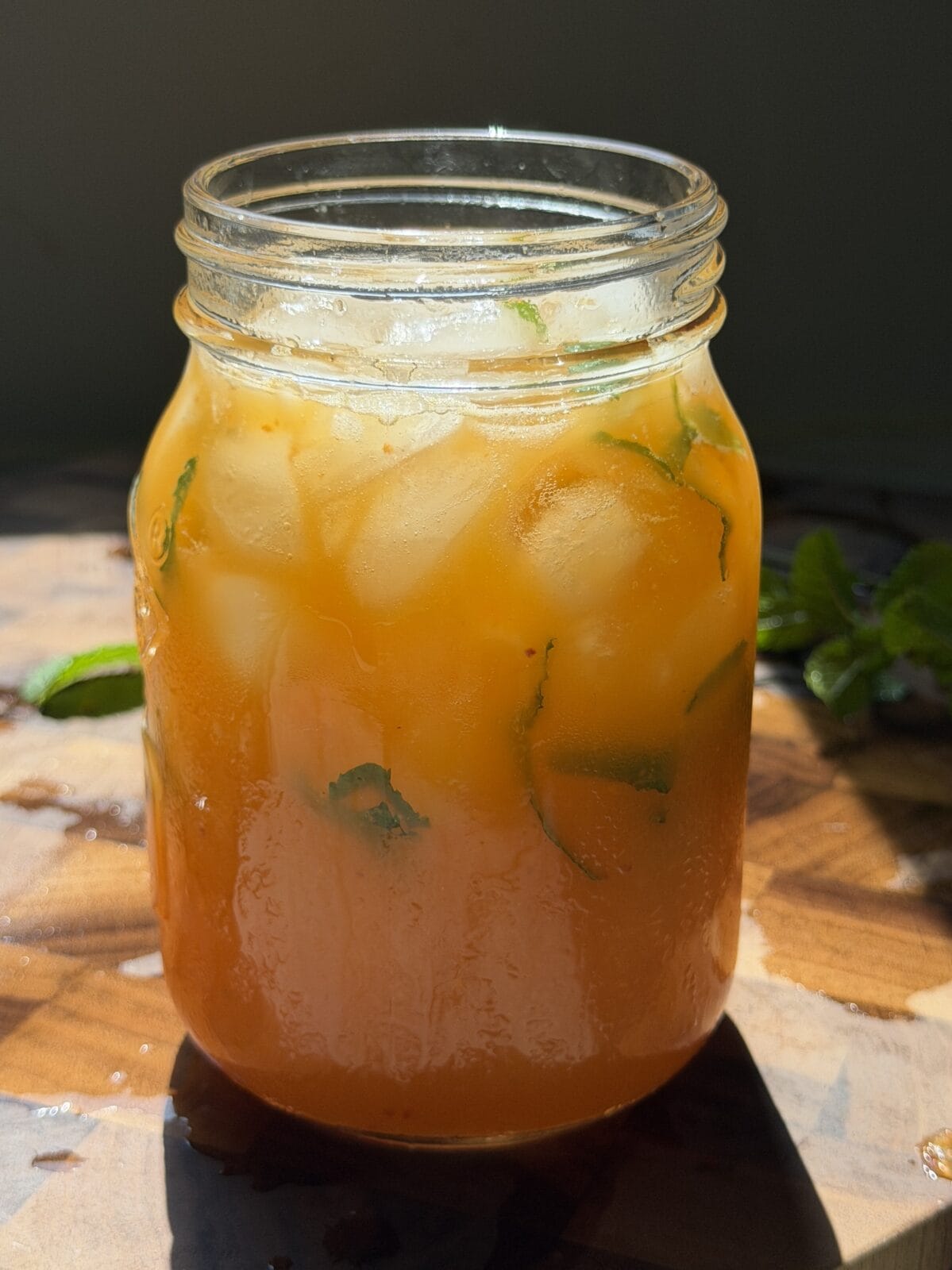 A mason jar filled with an orange iced drink containing mint leaves, sits on a wooden surface in bright sunlight.