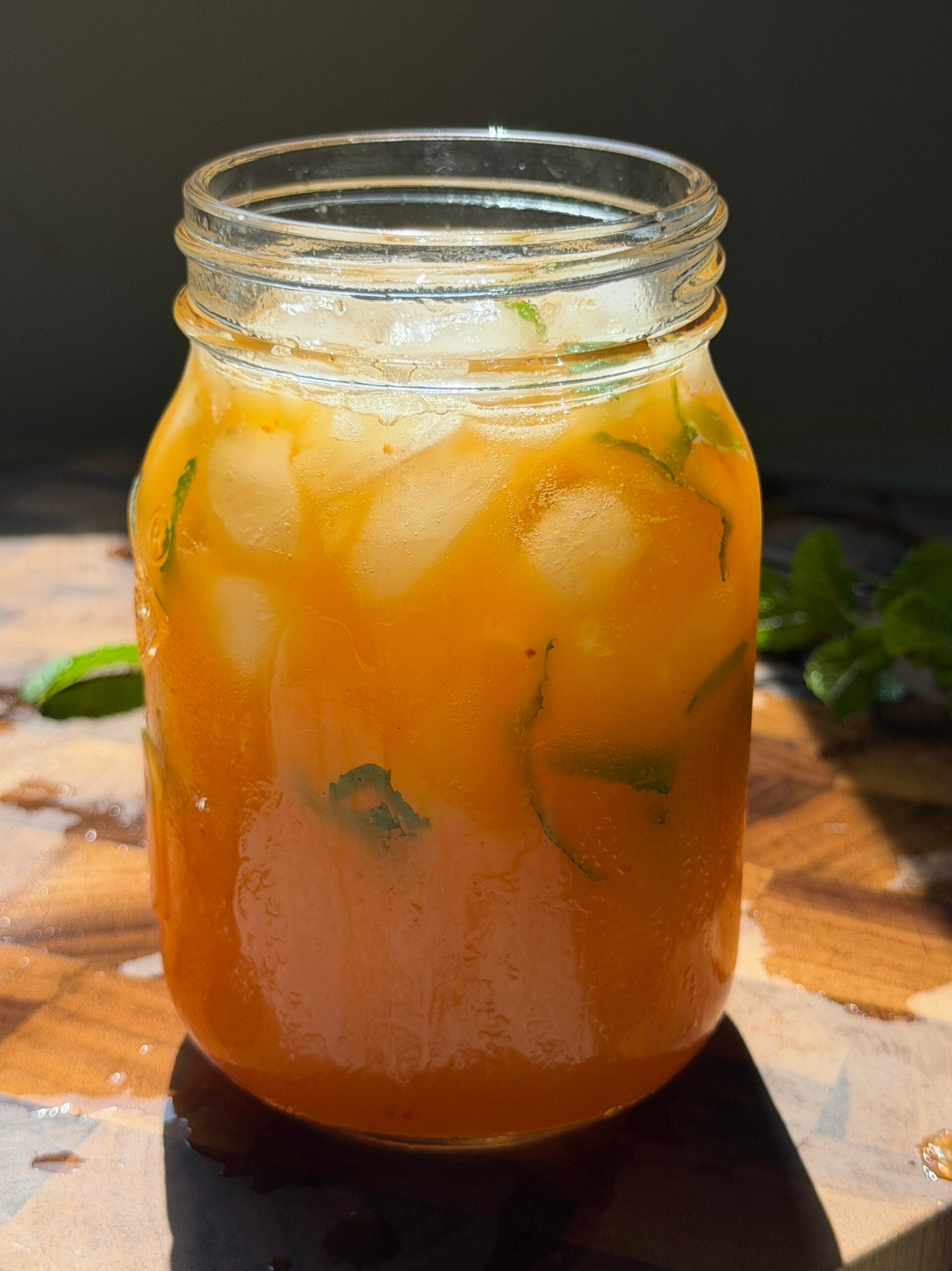 A mason jar filled with an orange iced drink containing mint leaves, sits on a wooden surface in bright sunlight.