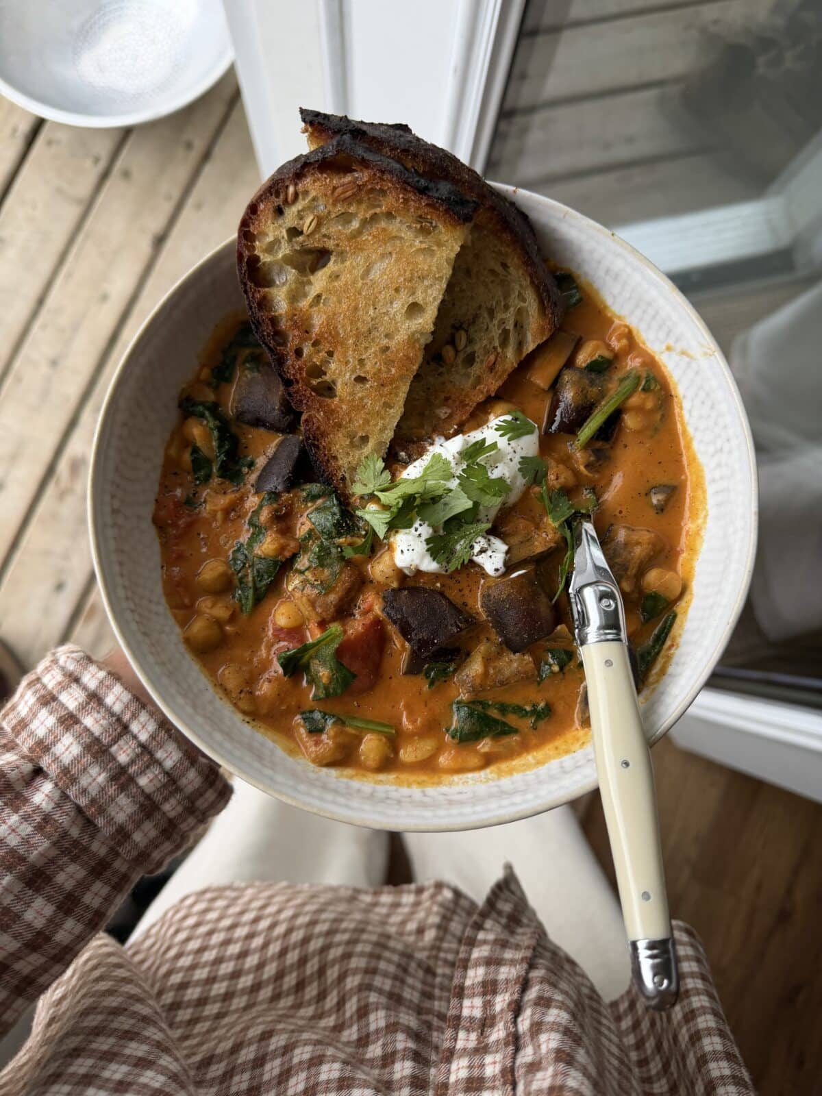 A person holds a bowl of vegetable stew topped with cilantro and sour cream, served with toasted bread; a fork rests on the bowl.