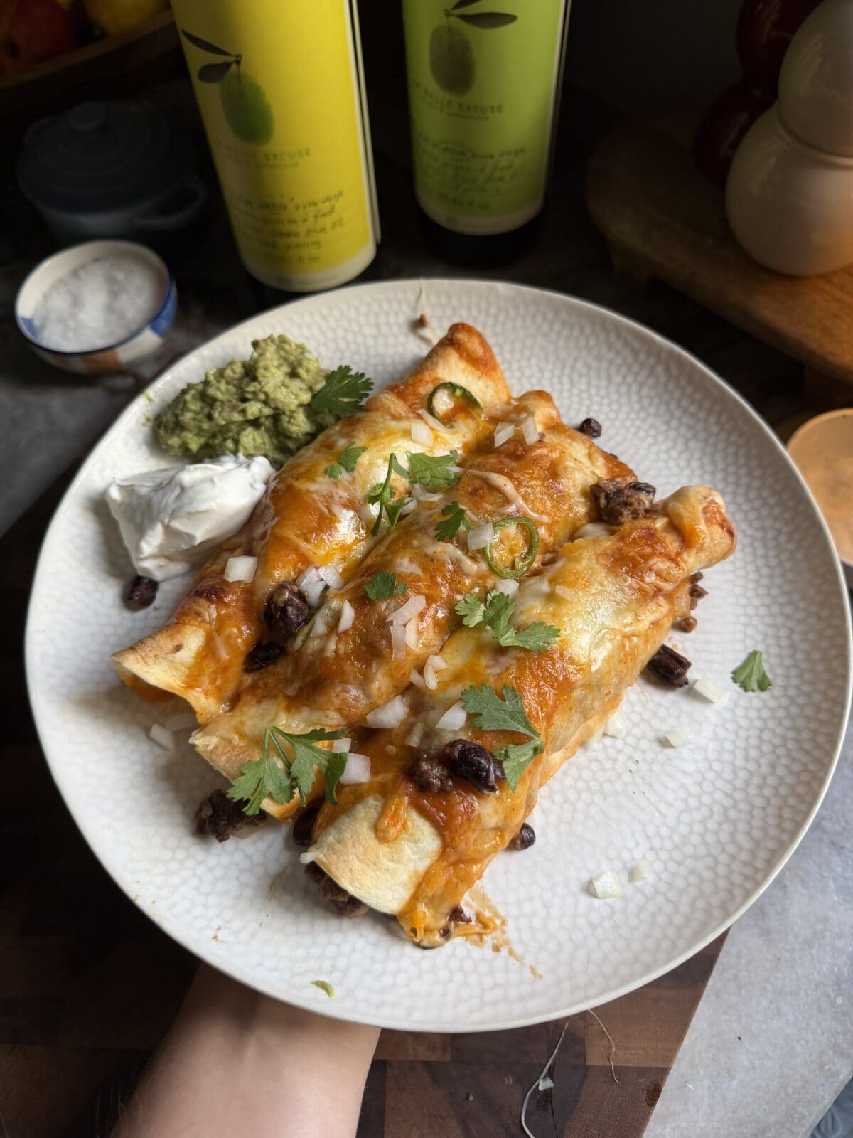 Three cheesy enchiladas topped with chopped onions and cilantro, served with sour cream and guacamole on a white plate.