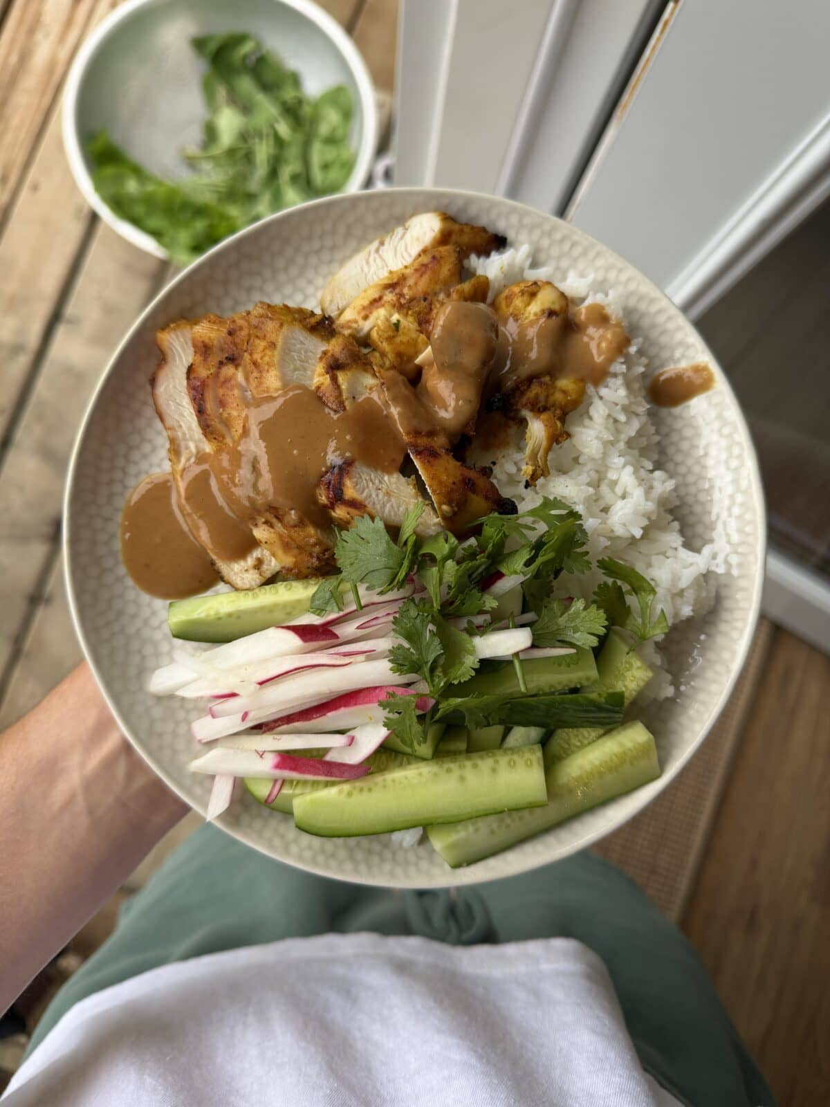 A bowl with grilled chicken, white rice, peanut sauce, sliced radish, cucumber, and cilantro is held over a wooden deck, with greens in a bowl in the background.