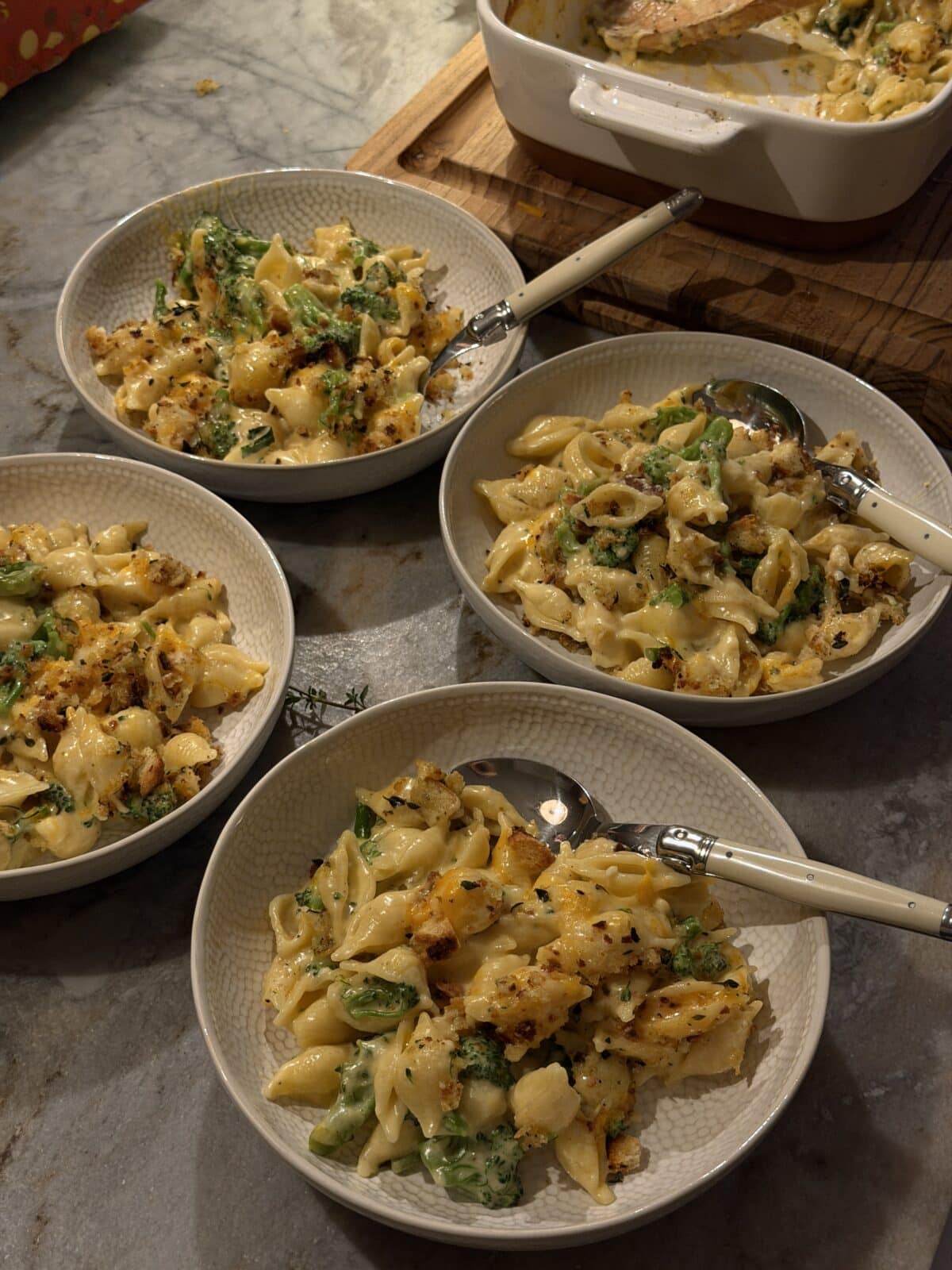 Three bowls of broccoli mac and cheese topped with breadcrumbs, each with a spoon, next to a baking dish on a counter.