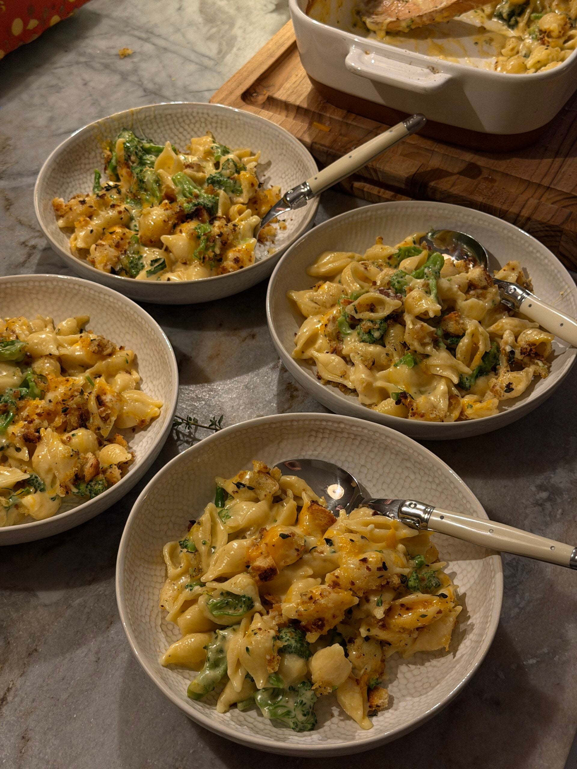 Three bowls of broccoli mac and cheese topped with breadcrumbs, each with a spoon, next to a baking dish on a counter.
