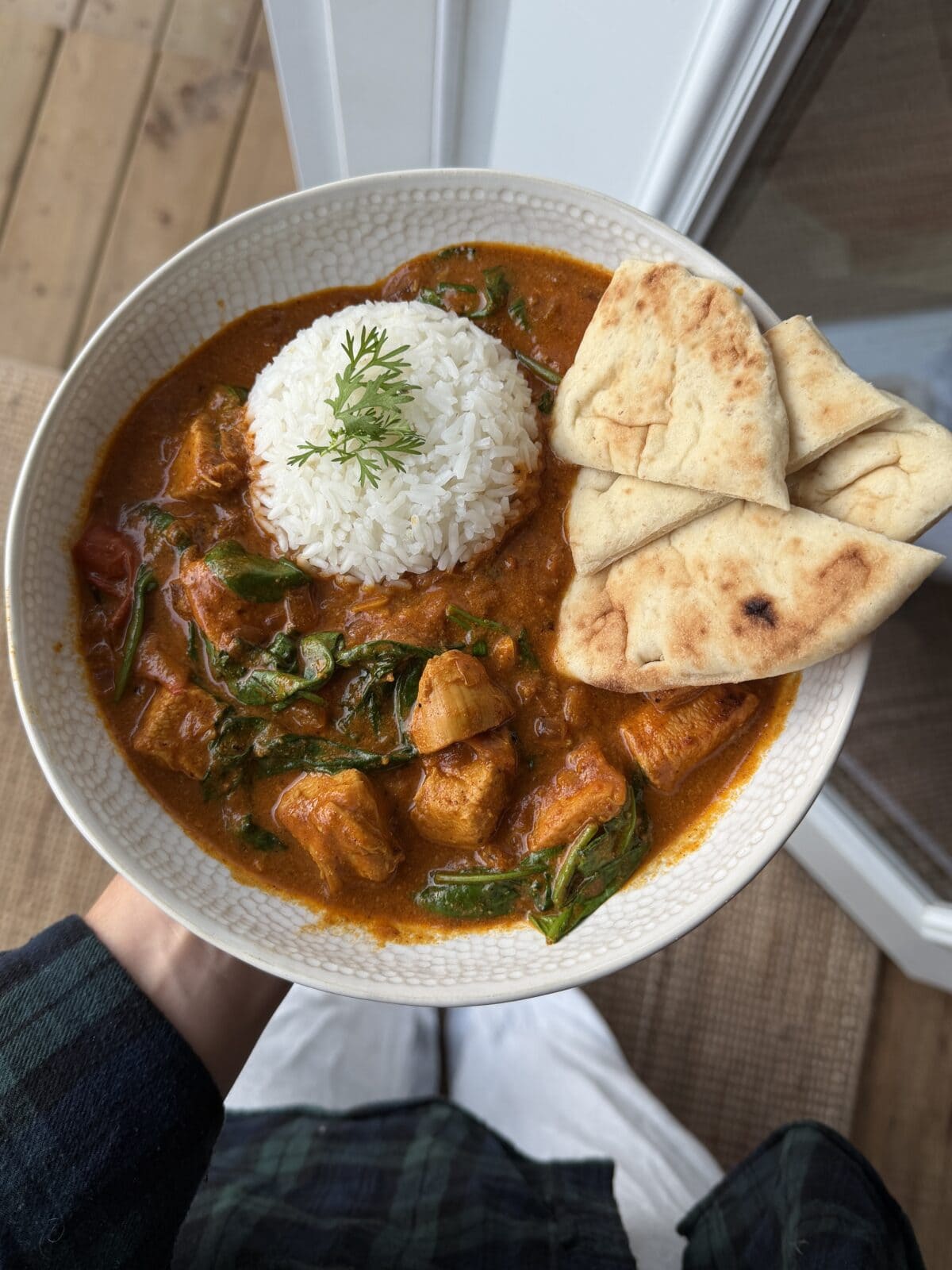 Bowl of curry with chunks of meat, white rice garnished with dill, wilted greens, and naan bread on the side, held by a person near a window.