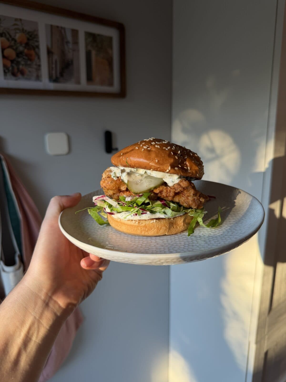 A hand holds a plate with a fried chicken sandwich topped with pickles and slaw on a sesame seed bun.