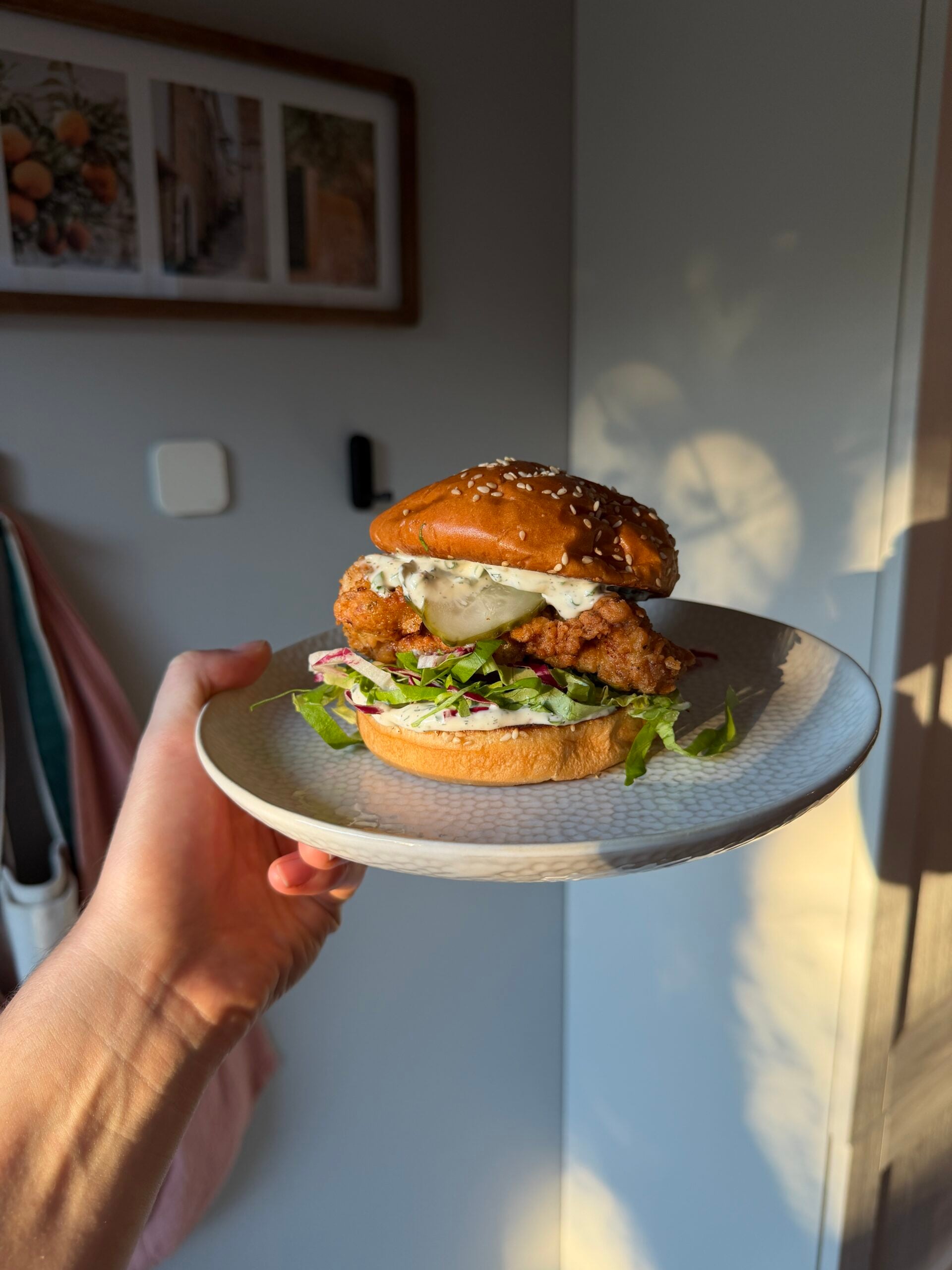 A hand holds a plate with a fried chicken sandwich topped with pickles and slaw on a sesame seed bun.