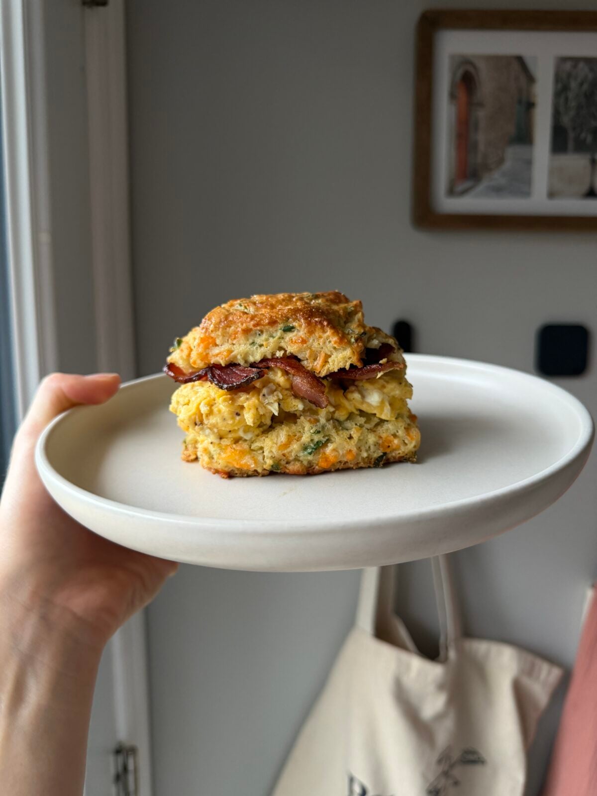 A hand holds a white plate featuring a biscuit breakfast sandwich with egg and bacon.