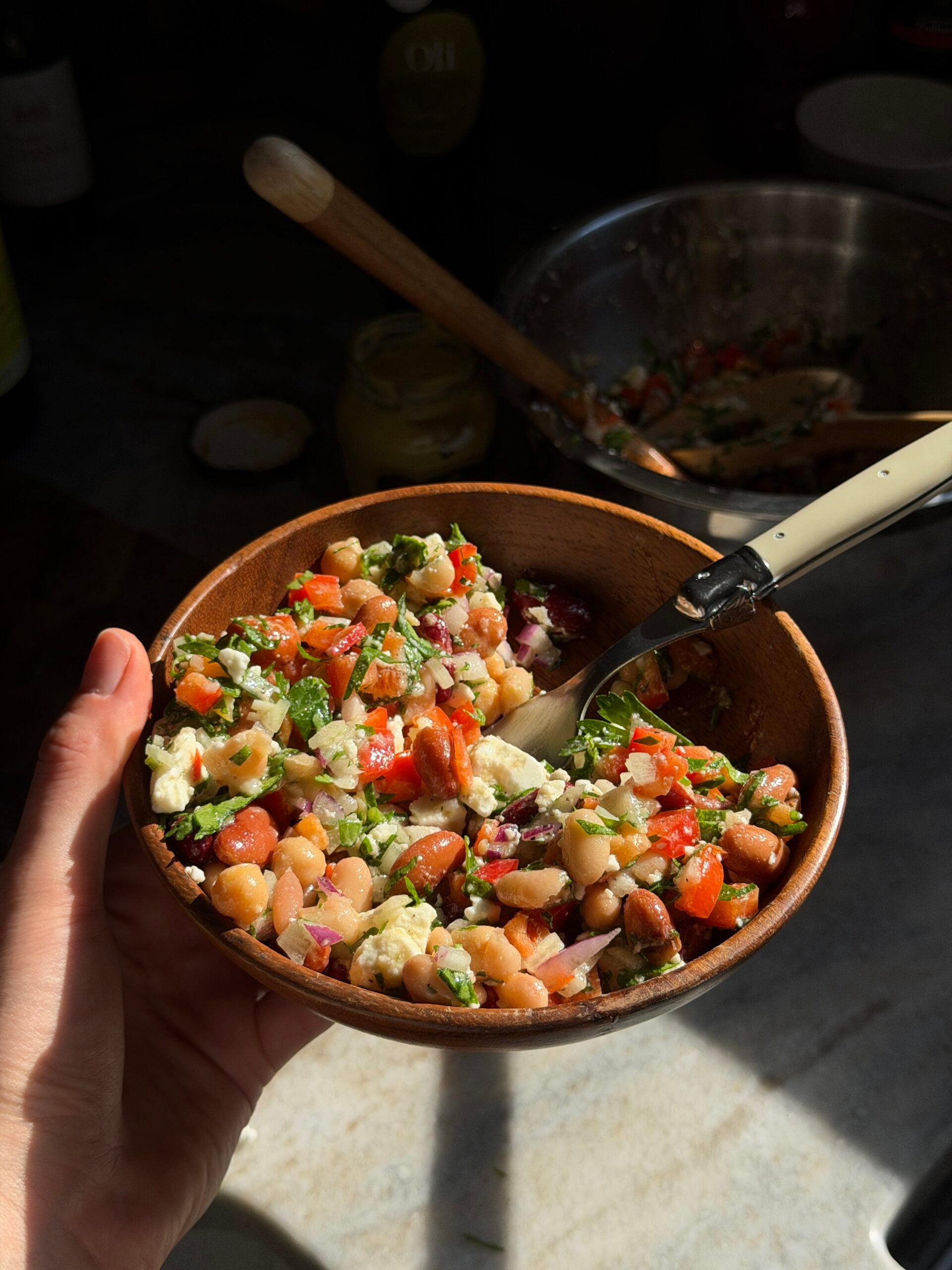 A hand holds a wooden bowl of bean and vegetable salad with crumbled cheese and a fork.