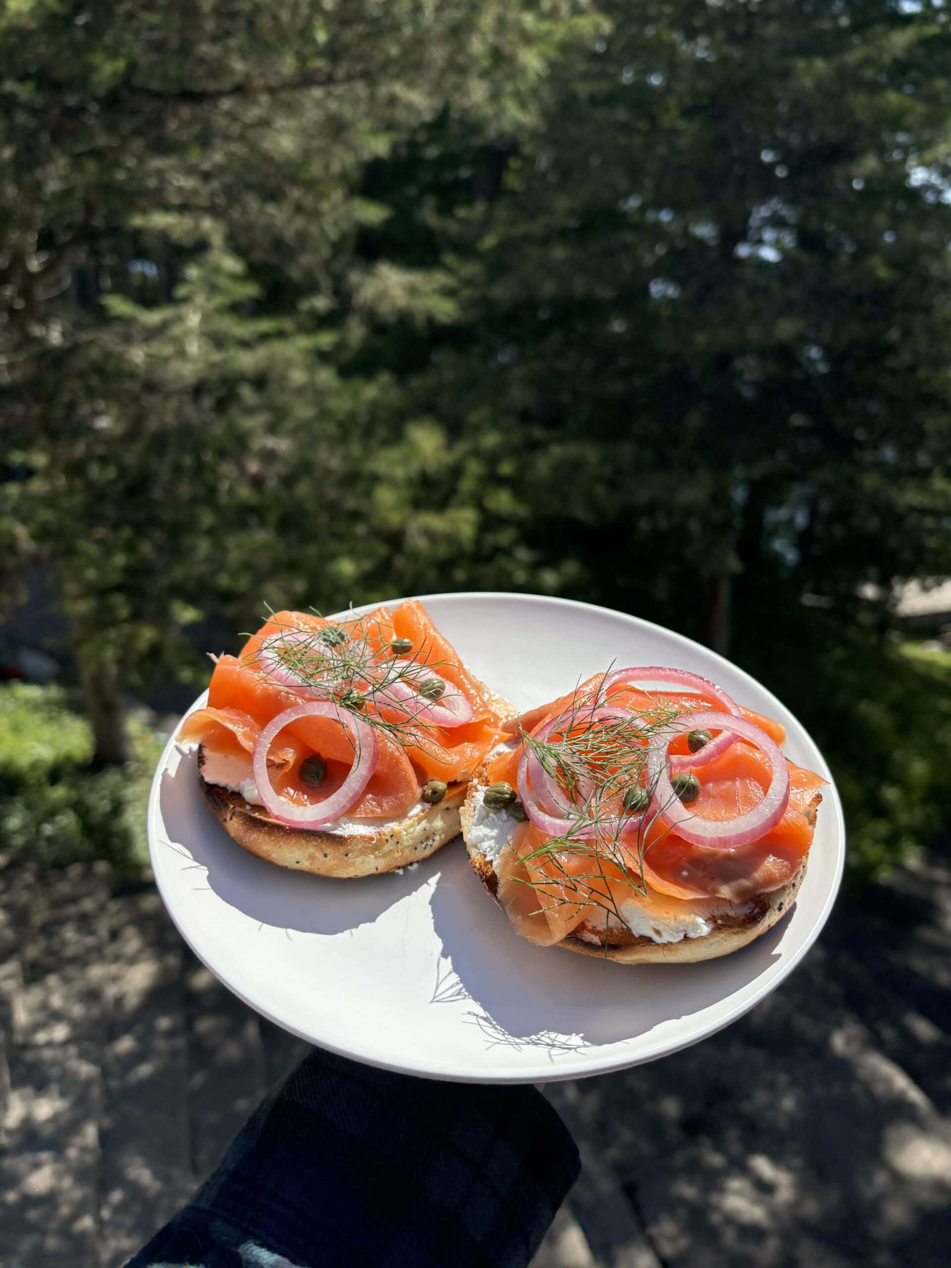 A white plate with two bagel halves topped with cream cheese, smoked salmon, capers, red onion slices, and dill, held outdoors with trees in the background.