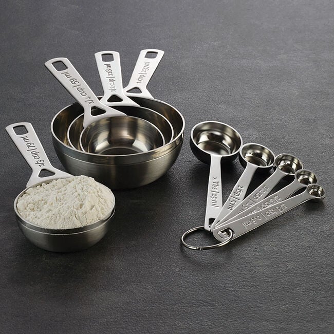 A set of metal measuring cups and spoons on a dark surface, with one cup filled with flour.