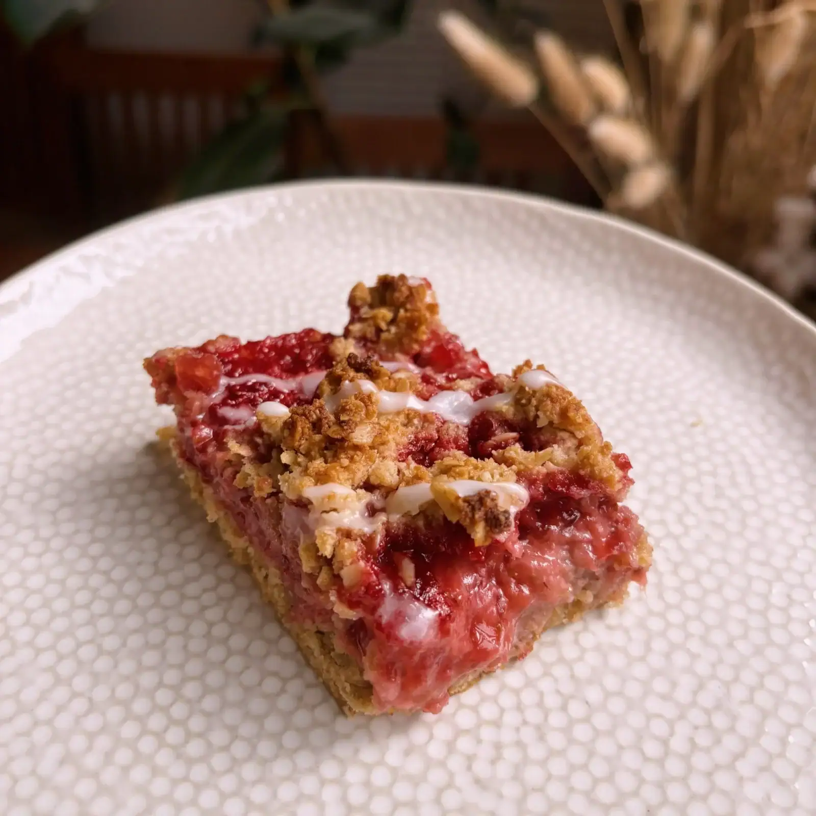 A close-up of a single raspberry crumb bar with icing, served on a textured white plate.