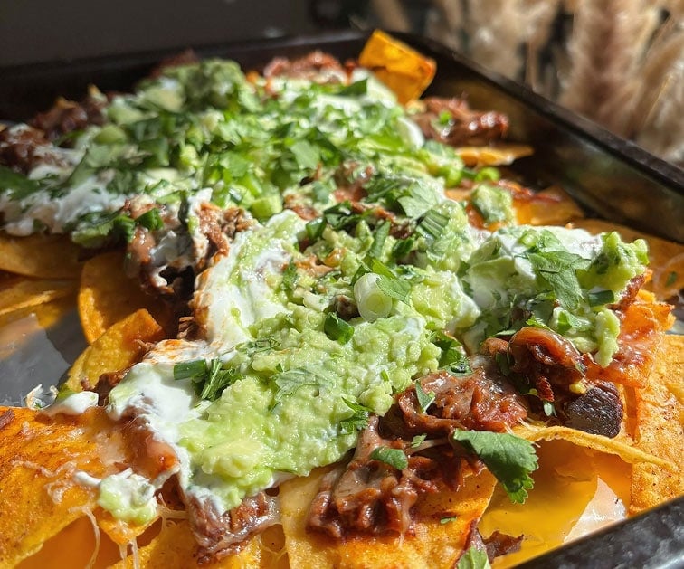 A close-up of nachos topped with shredded meat, guacamole, sour cream, chopped cilantro, and melted cheese on a tray.