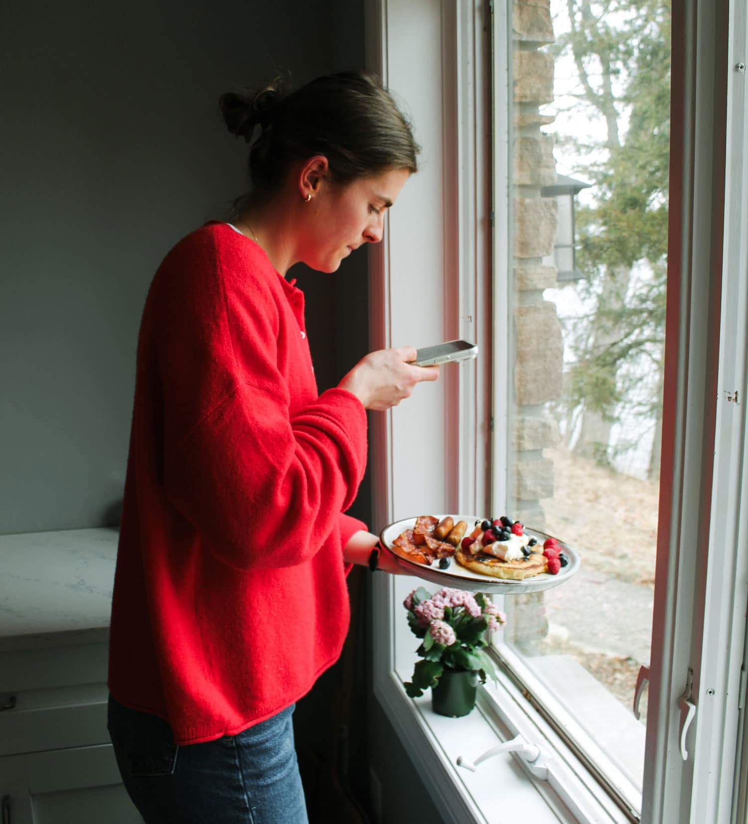 A woman in a red sweater takes a photo of a plate of waffles with fruit by a window.