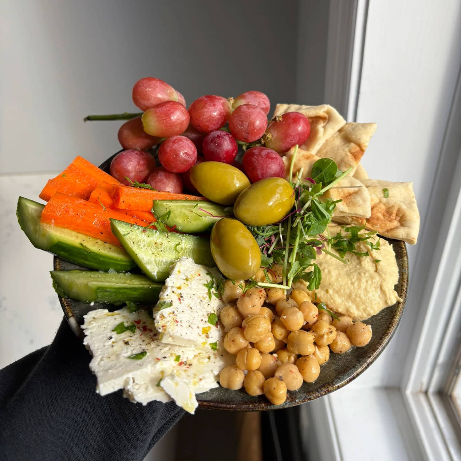 A bowl containing grapes, pita, olives, hummus, chickpeas, feta cheese, cucumber, carrot sticks, and microgreens held by a person near a window.