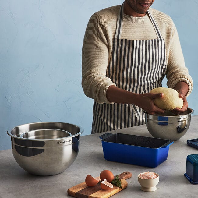 Person wearing a striped apron kneads dough over a metal bowl on a counter with eggshells, a blue baking dish, a small bowl of flour, and herbs.