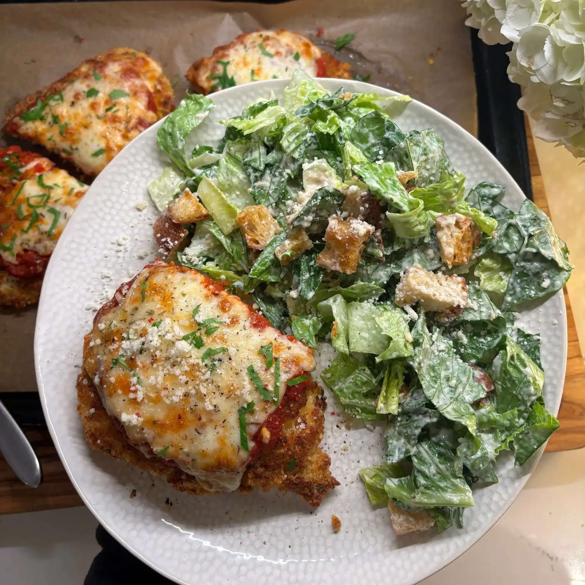 Plate with chicken parmesan topped with melted cheese and herbs, served with Caesar salad and croutons. More chicken parmesan is visible on a baking tray in the background.