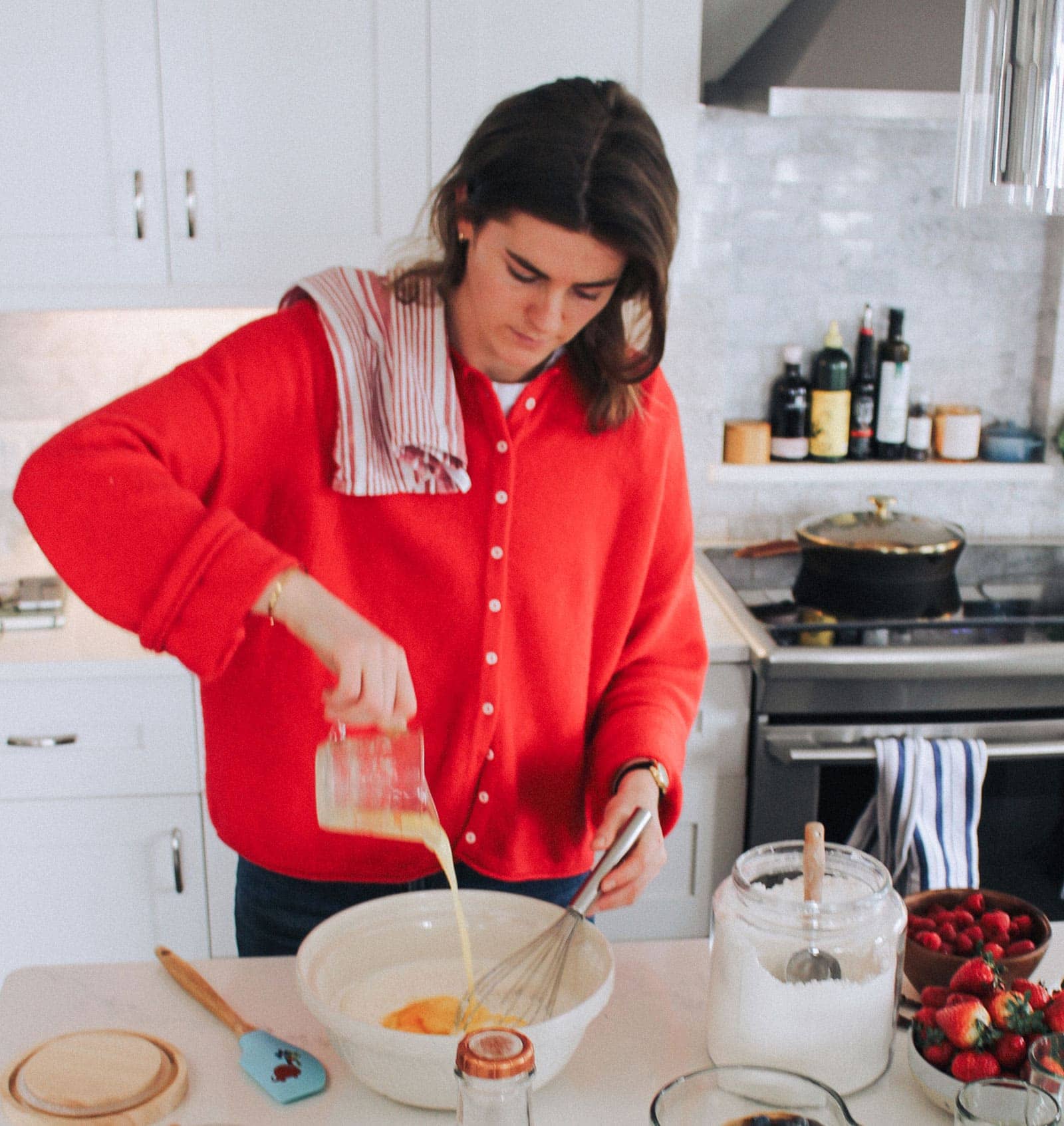 A person in a red sweater pours liquid into a mixing bowl in a kitchen, with baking ingredients and utensils on the counter.